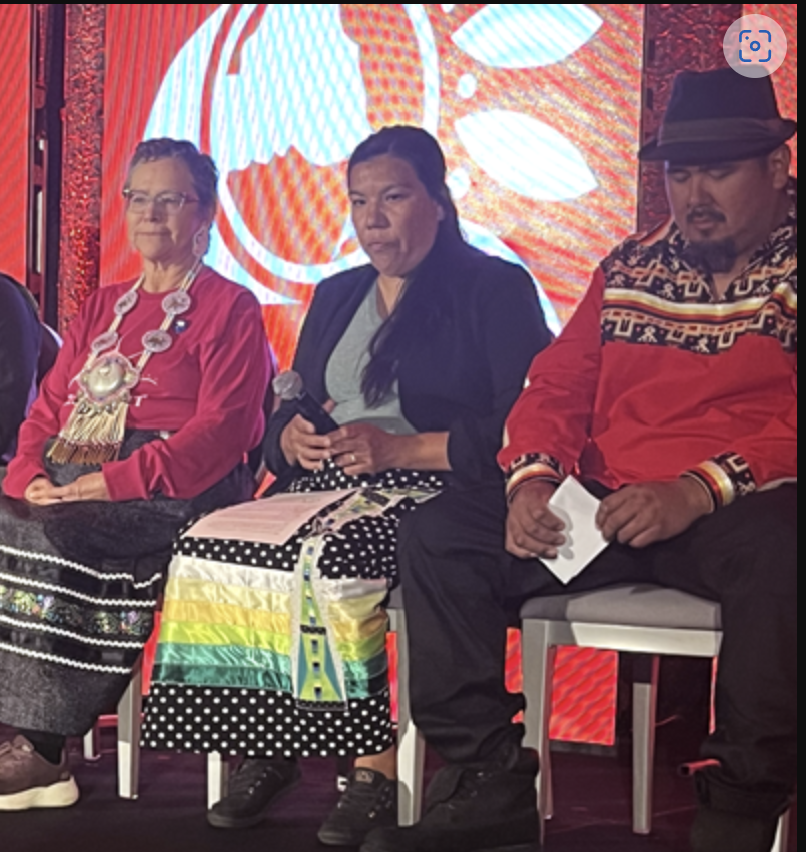 Three individuals seated on a stage, holding microphones and papers, with a large red and white graphic backdrop, and a rainbow flag on a cloth in front of the middle person.