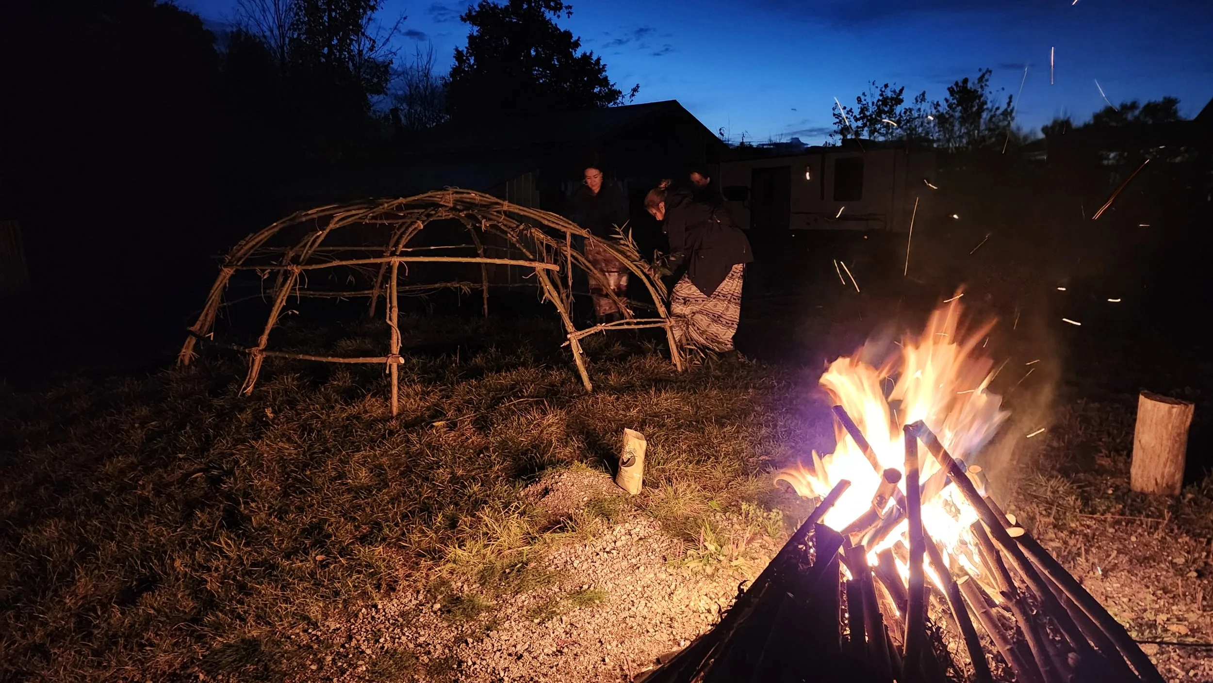 People gathered around a campfire at night, constructing a dome-shaped shelter with wooden sticks.