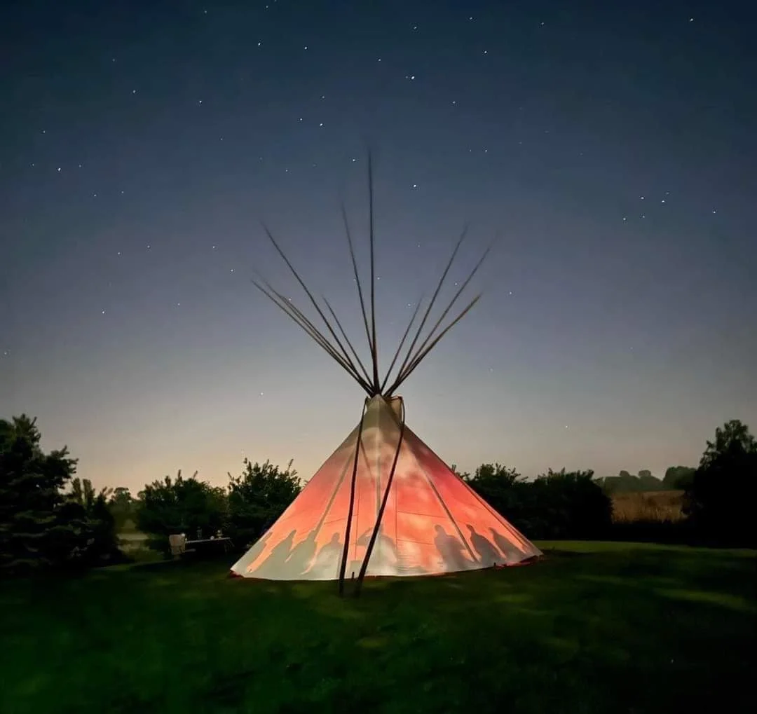A tipi tent illuminated with a red-orange light on a grassy area at night, with stars visible in the sky and trees in the background.