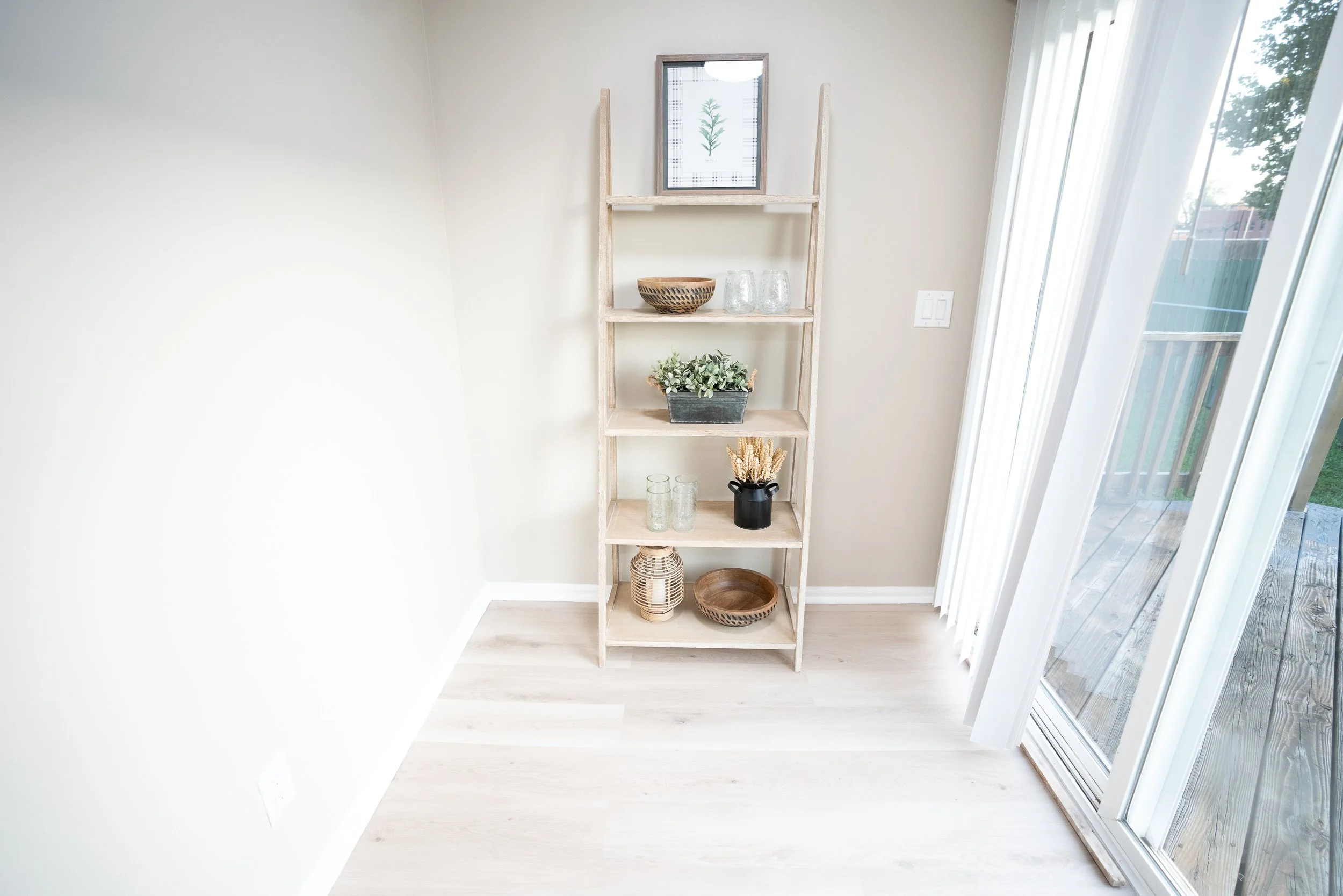 Bright room with a rustic ladder-style shelf displaying decorative bowls, glass jars, plants, and framed botanical art beside a large window.