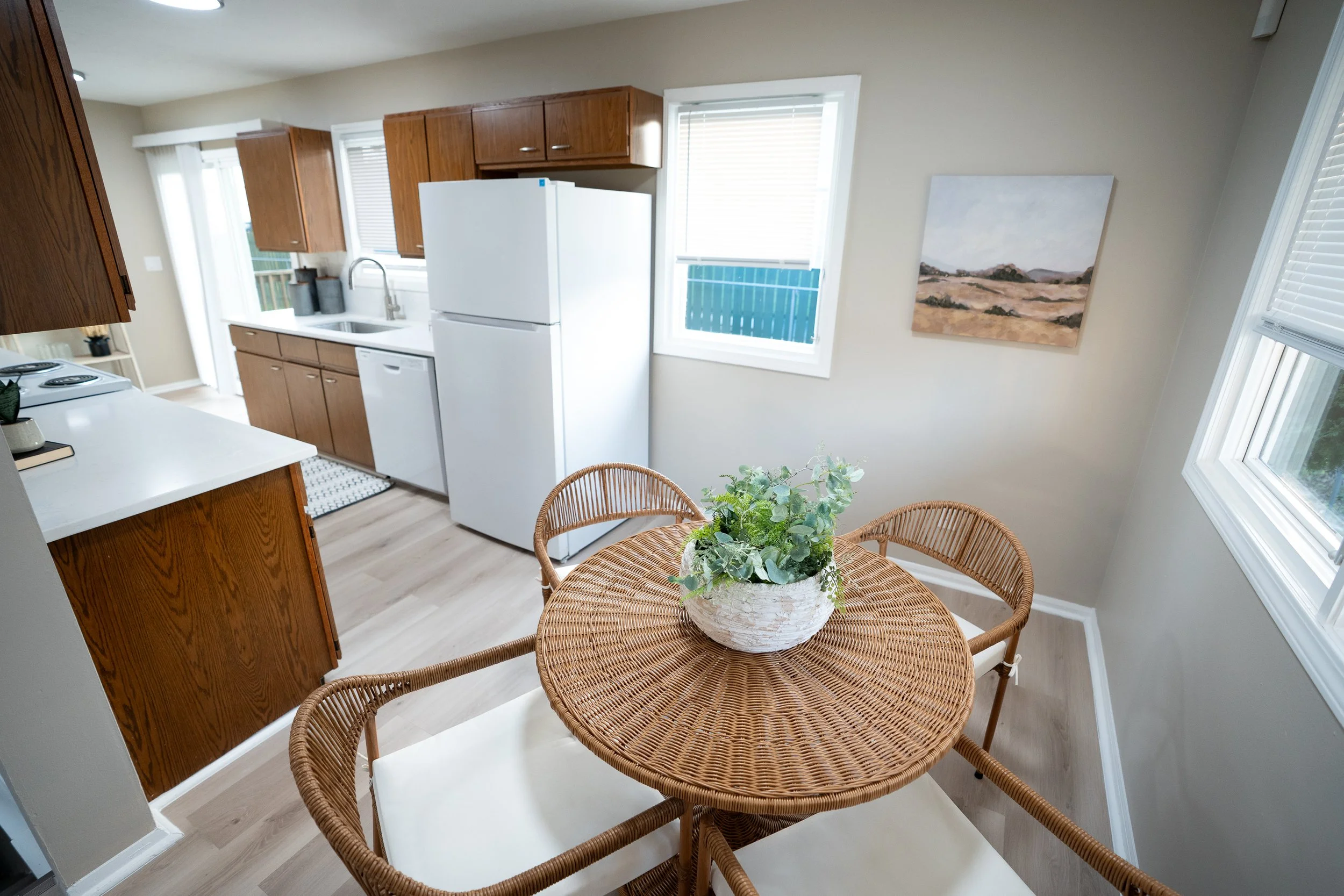 Bright kitchen dining area with wood cabinets, white appliances, wicker table and chairs, and a potted centerpiece