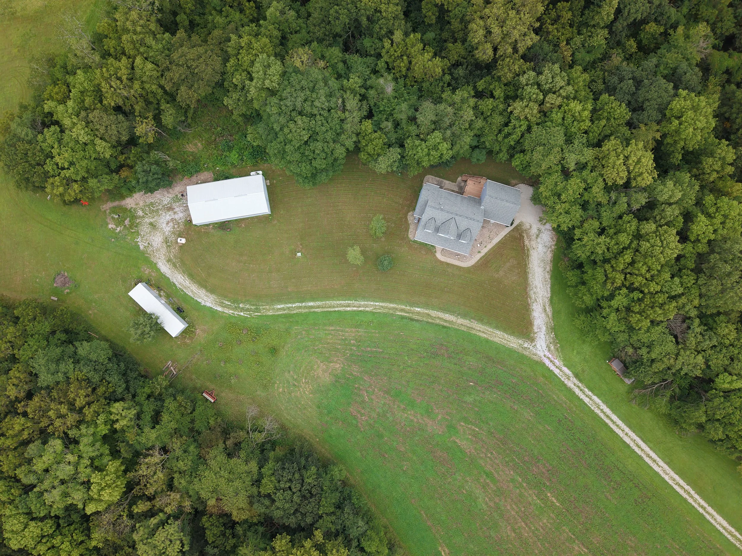 Aerial view of a rural property with a house, barns, gravel driveways, open lawn, and surrounding dense forest.