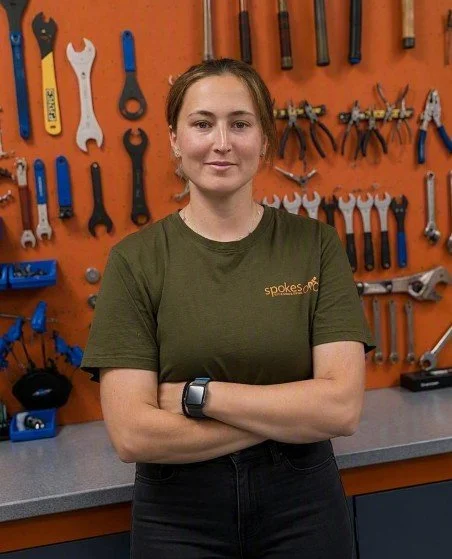 A woman with brown hair, wearing a green t-shirt and a smartwatch, standing with arms crossed in a workshop. The background has orange walls with various tools like wrenches, screwdrivers, and pliers hanging on them.