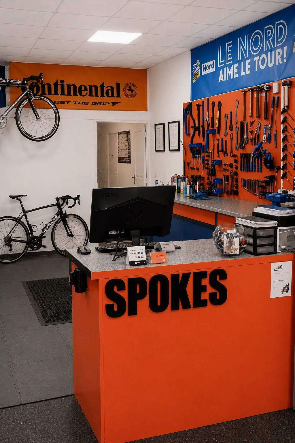 Bike shop reception area with orange and blue color scheme, featuring a counter labeled 'SPOKES,' a computer, a glass container with dog figurines, a wall with bike tools, and bicycles on display.