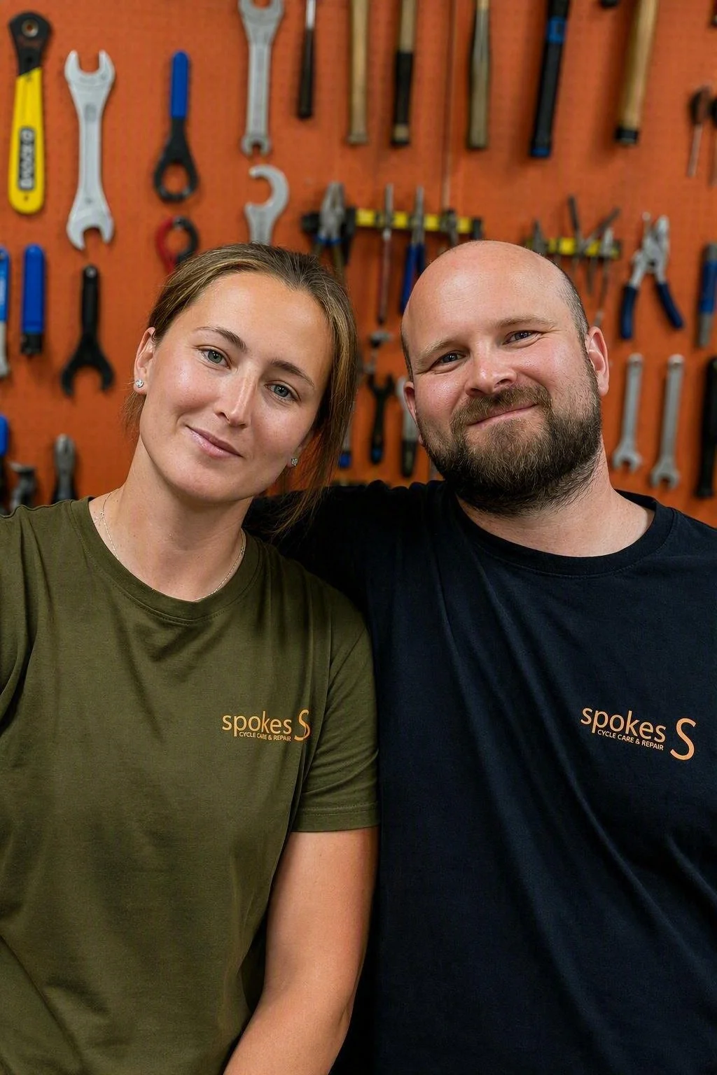 A man and a woman posing with their arms around each other in a workshop with a pegboard wall filled with tools behind them. Both are wearing shirts with the logo 'spokes' and the words 'Cycle Care & Repair'.