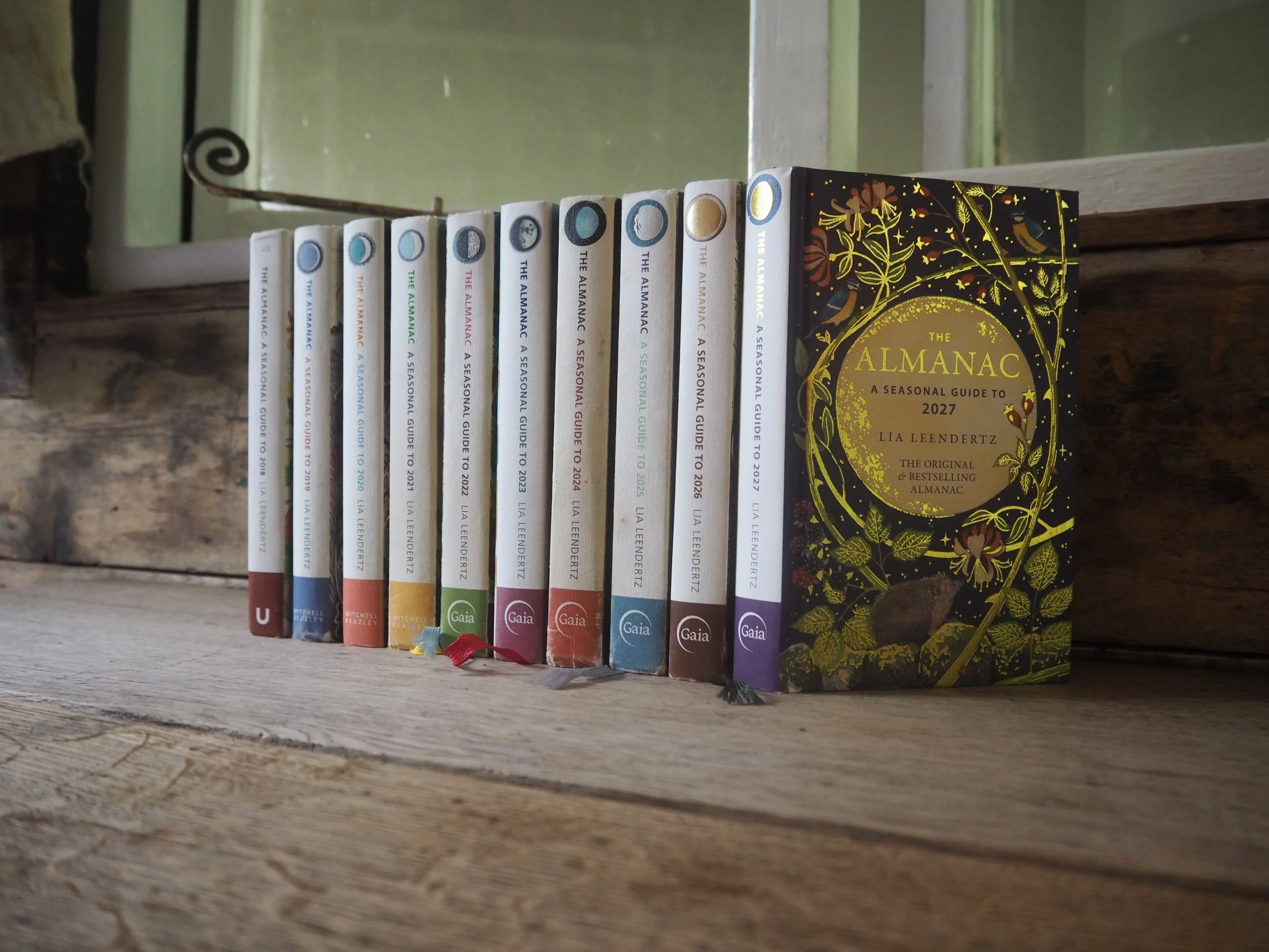 A row of colorful books titled 'The Almanac: A Seasonal Guide to 2027' by Lia Leendertz, standing on a wooden surface next to a mirror with a decorative scrollwork frame, with a rustic background.
