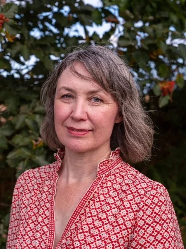 A woman with short, wavy, gray hair standing outdoors in front of green foliage. She is wearing a red and white patterned top and has a slight smile.
