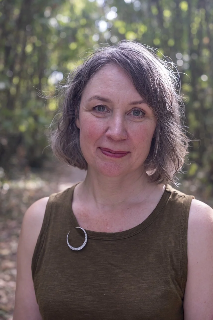 A woman with short, wavy gray hair standing outdoors in a wooded area, wearing a sleeveless brown top with a decorative brooch on the shoulder.