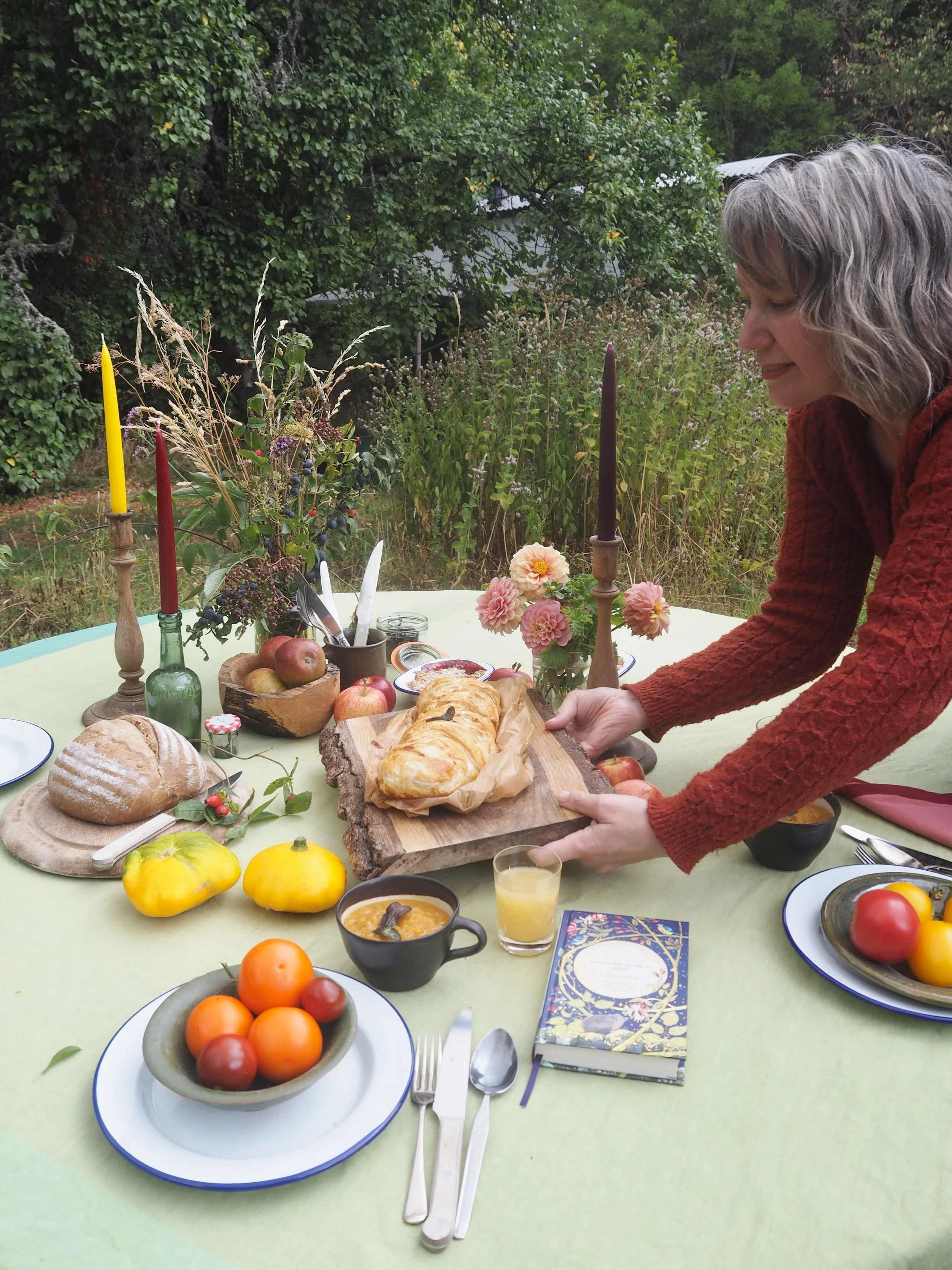 A woman preparing to serve a meal at an outdoor table decorated with autumn-themed candles, flowers, and a variety of food including bread, apples, tomatoes, and pumpkin desserts, with greenery and trees in the background.