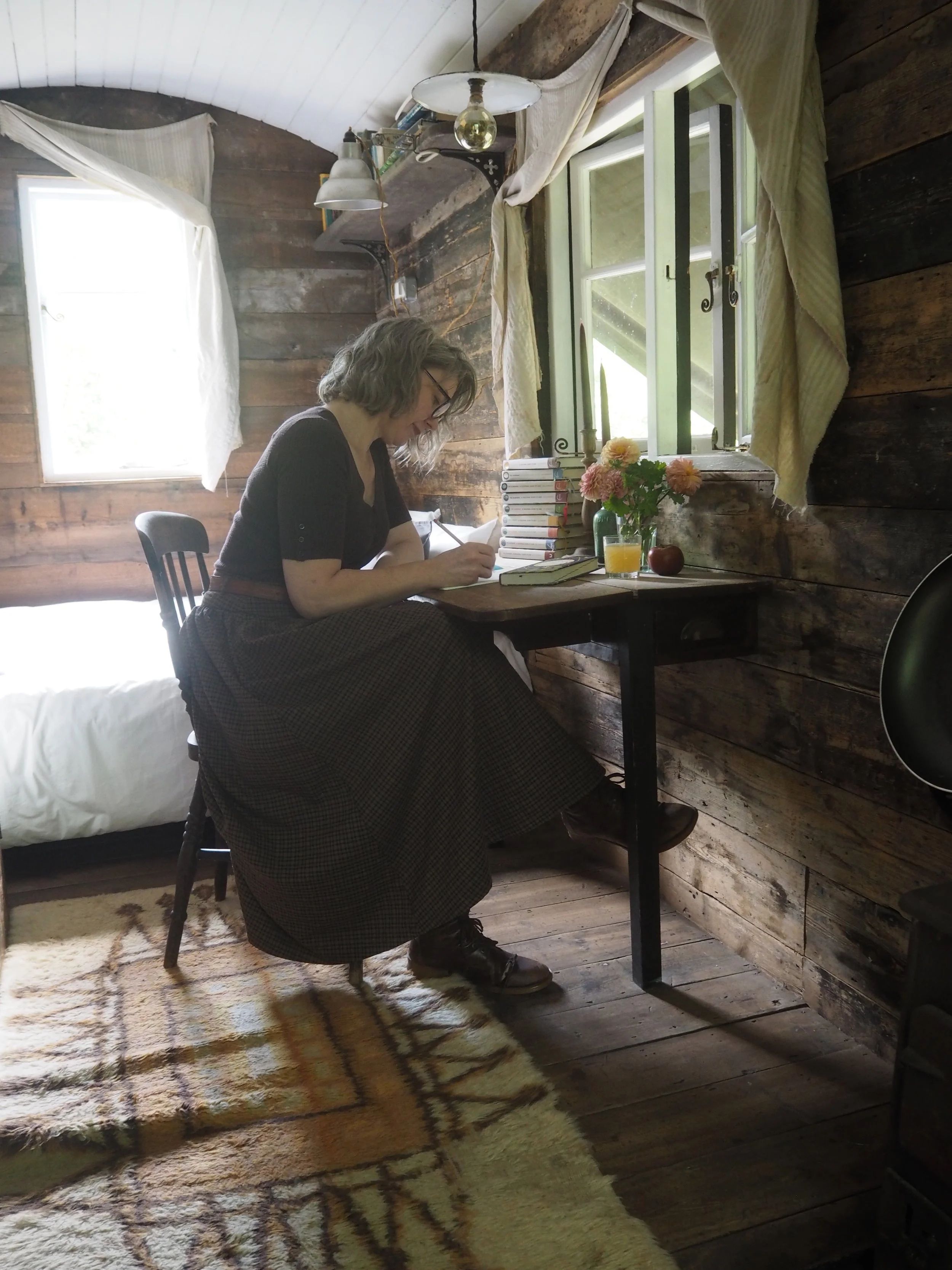 A woman with shoulder-length hair, glasses, and wearing a brown sweater, is sitting at a wooden desk writing. The desk has a stack of books, a bouquet of pink flowers in a green vase, a glass of orange juice, and an apple. The room has wooden walls, two windows with curtains, a ceiling lamp, and a bed with white bedding. The floor is wooden with a beige and orange patterned rug.