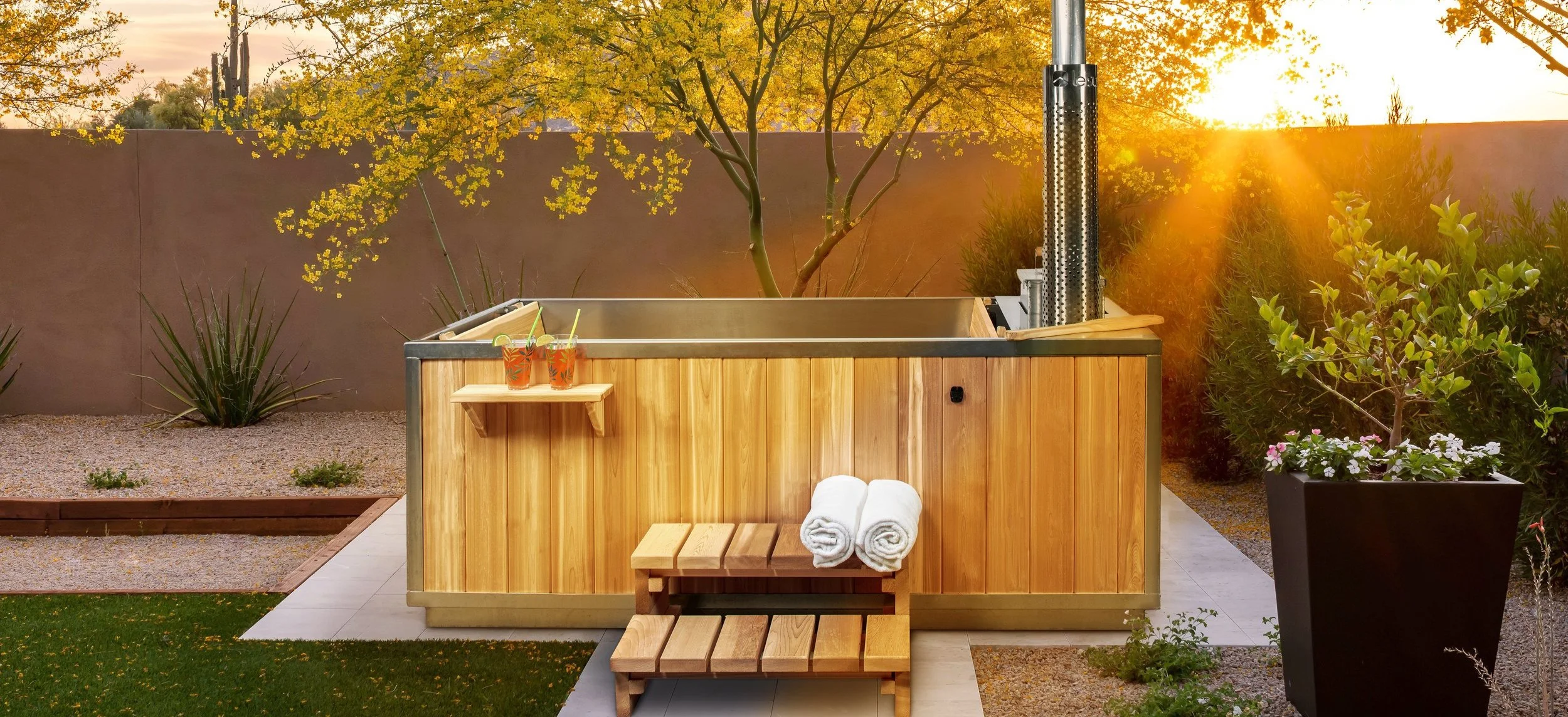 An outdoor hot tub with a wooden exterior in a landscaped backyard during sunset. Two drinks with lime and straws are placed on a small wooden shelf attached to the hot tub. Towels are rolled and placed on a small bench below the shelf. There are trees, plants, and a tall wall in the background.