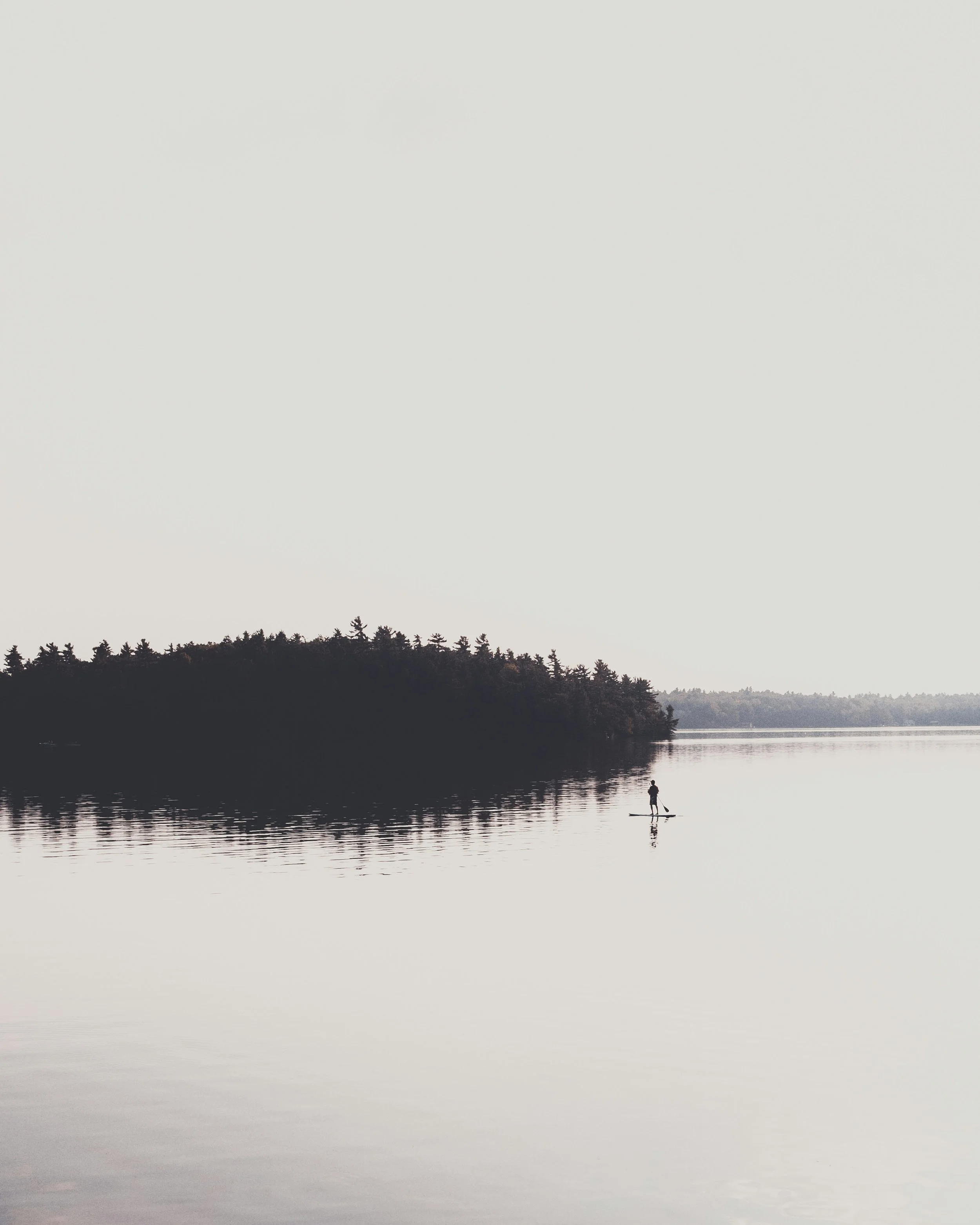 A person paddleboarding on a calm lake with a shoreline lined with trees in the distance under an overcast sky.
