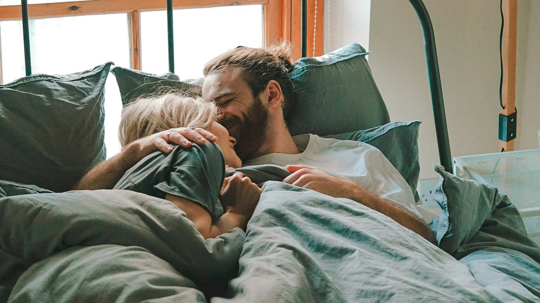 A couple sharing an intimate moment in bed, smiling and touching foreheads, with pillows and a bed frame around them.