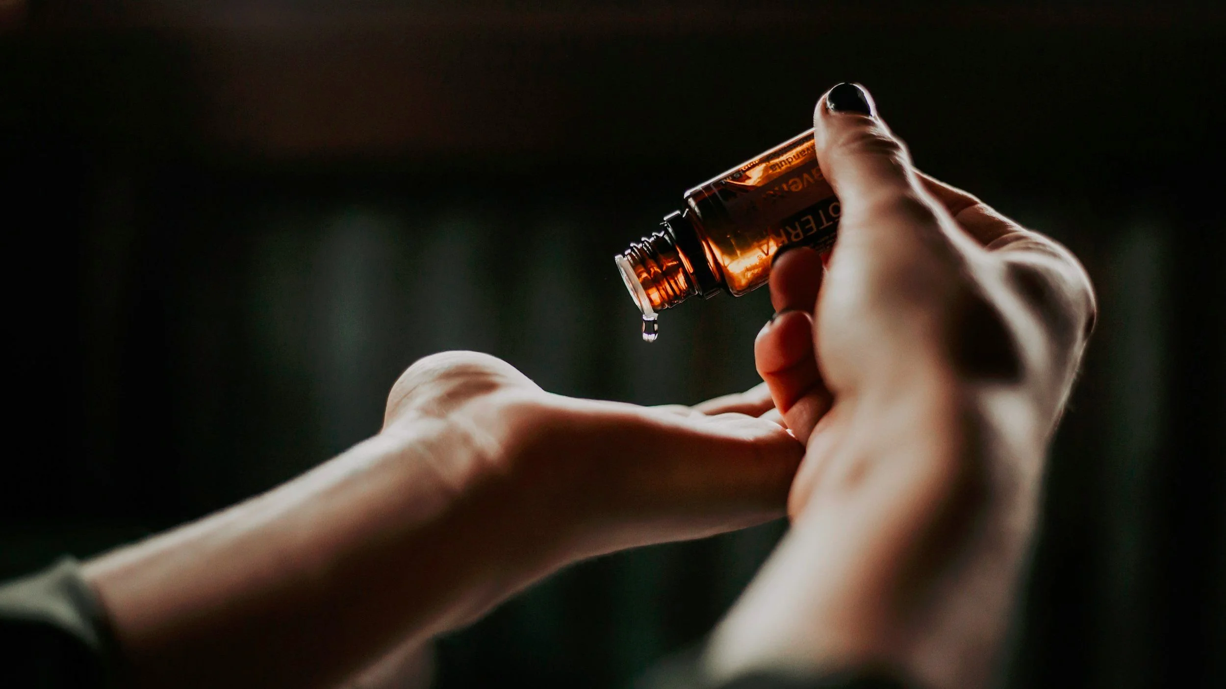 Person holding a small glass vial or bottle with liquid, with a drop falling from it, in a dark setting.