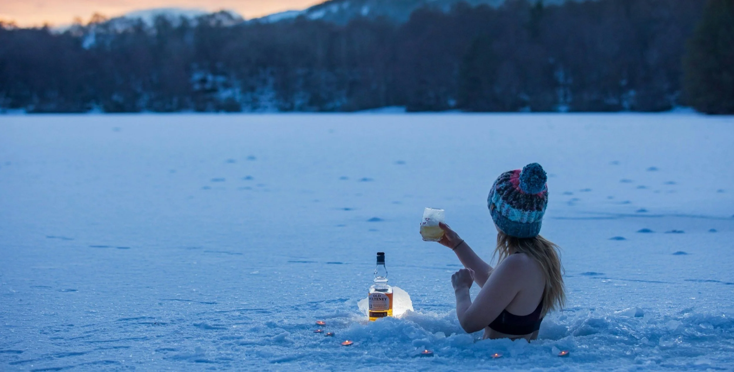 A person with long hair wearing a colorful knit beanie is sitting in the snow by a frozen lake during dusk or dawn, holding a glass of a beverage, with a bottle of liquor in front of them. Small candles are placed around in the snow for ambiance.