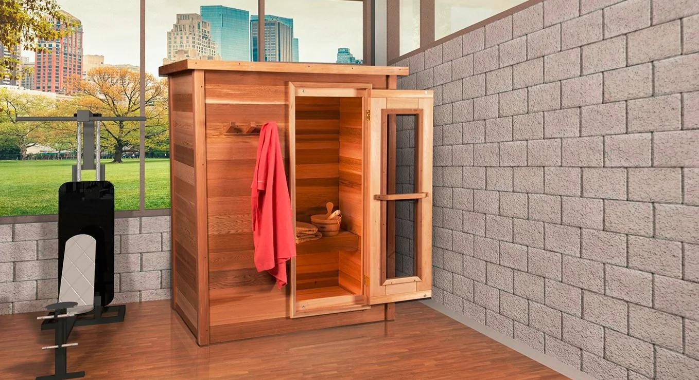 An outdoor sauna with a glass door, situated on a wooden deck. There is a towel hanging on a hook outside the sauna. To the left of the sauna, there is an exercise bench with weights. The background features a cityscape with tall buildings and a park with trees.