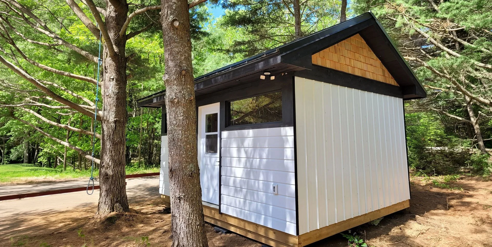A small, modern shed with black trim, white siding, and a wooden section under the roof, situated in a wooded area with trees around.