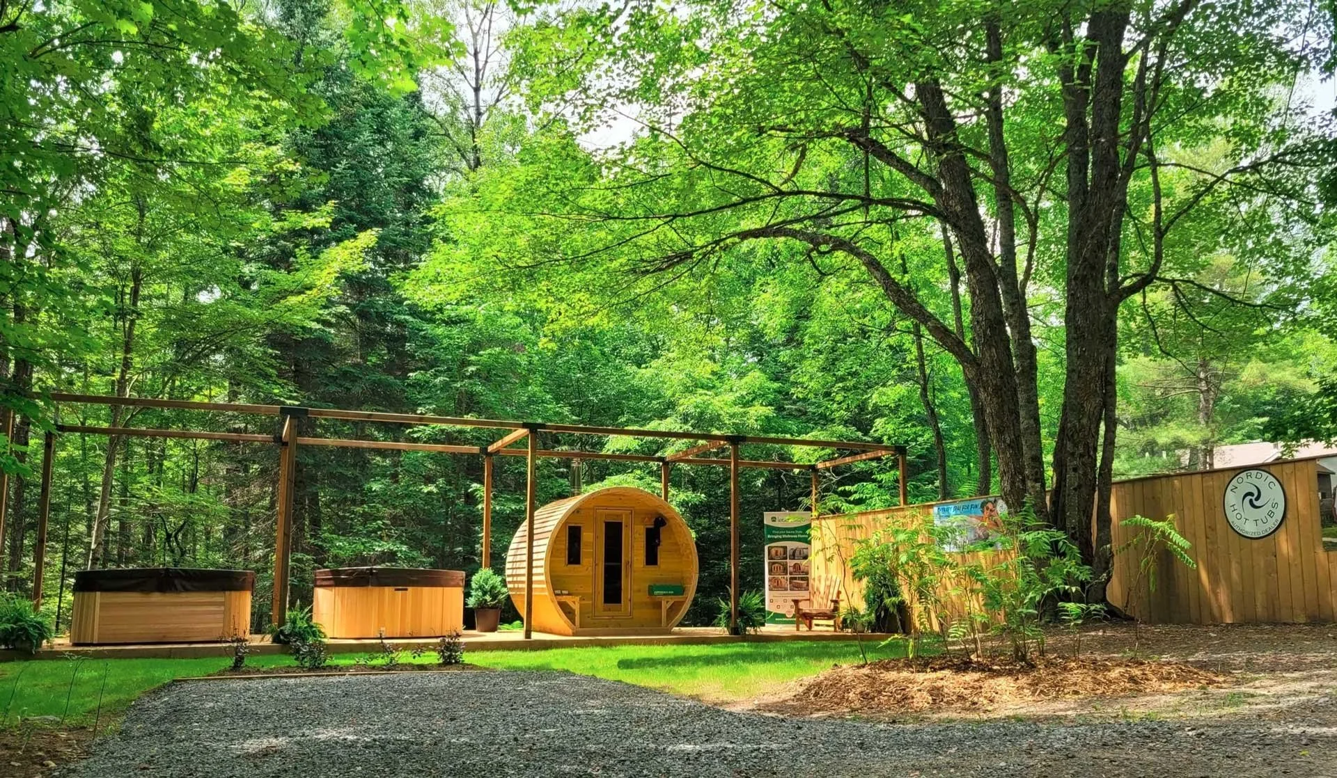 A small wooden sauna cabin surrounded by trees in a lush green forest, with a gravel path leading past it.