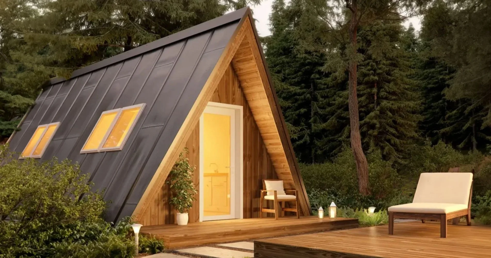 A small A-frame cabin with a black metal roof and yellow window panes, surrounded by trees and green foliage, featuring a porch with outdoor furniture and candles at dusk or evening.