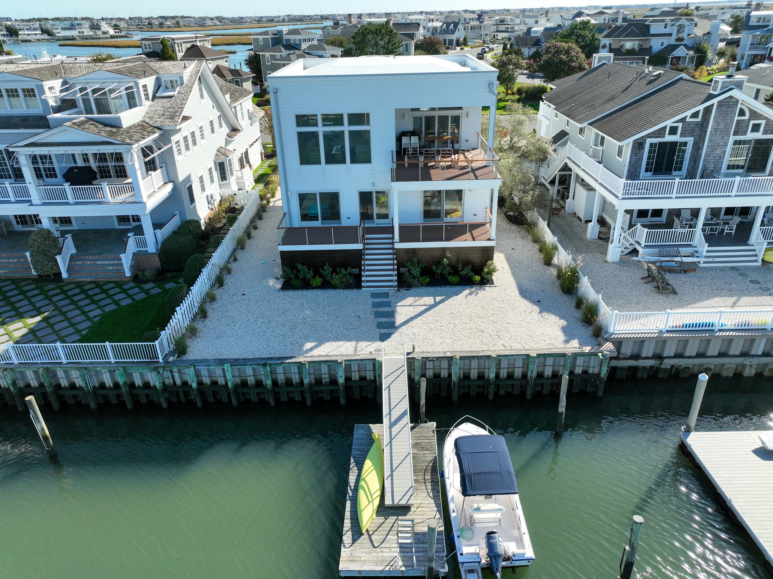 Aerial view of a white modern house on the bay with a boat docked at a pier, surrounded by similar houses and a marina in the background.