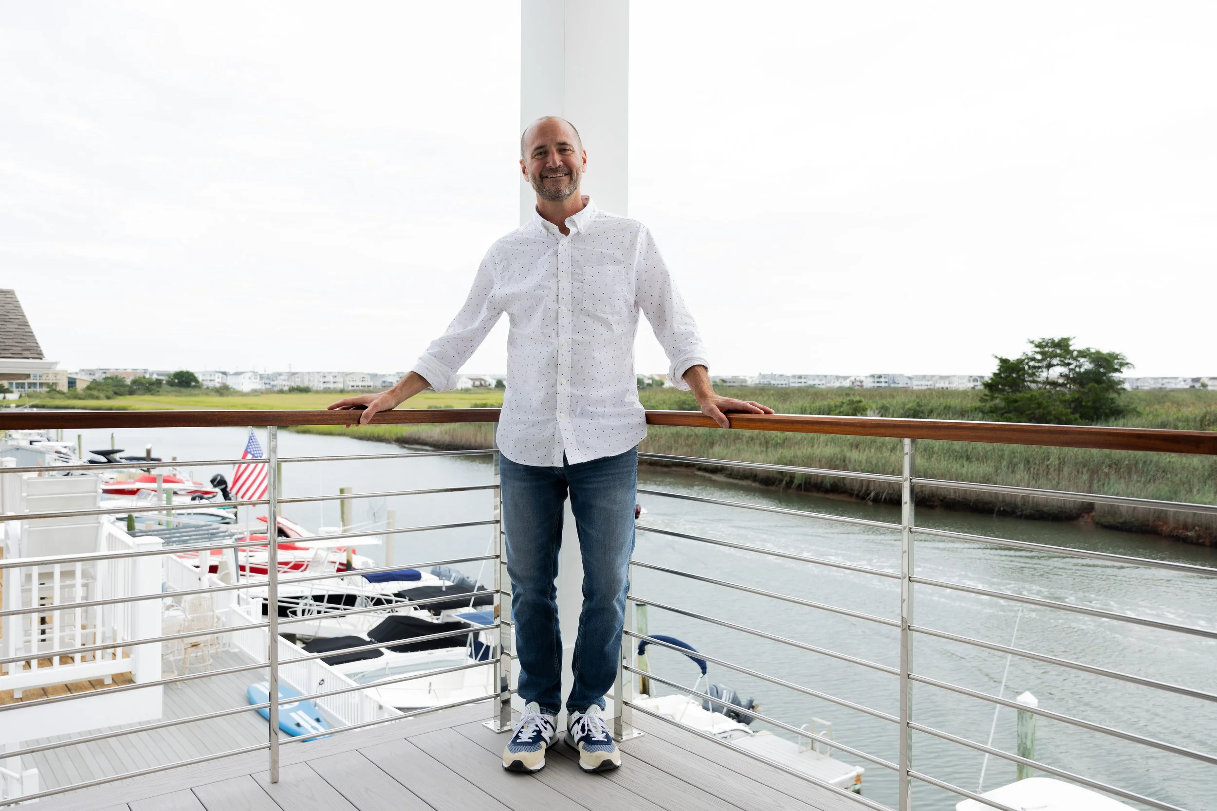Man smiling on a balcony overlooking water and boats with houses in the background.