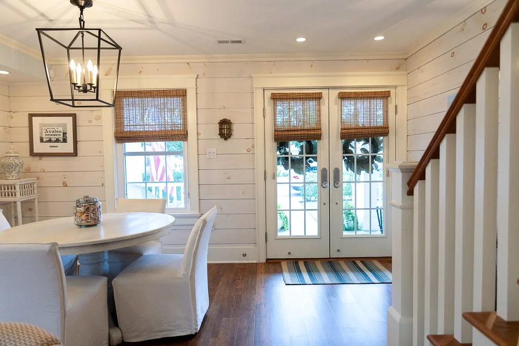 Interior of a cozy dining area with white painted wooden walls, a round white table surrounded by four white slipcovered chairs, a lantern-style chandelier hanging from the ceiling, and a set of French doors with bamboo Roman shades leading outside. 