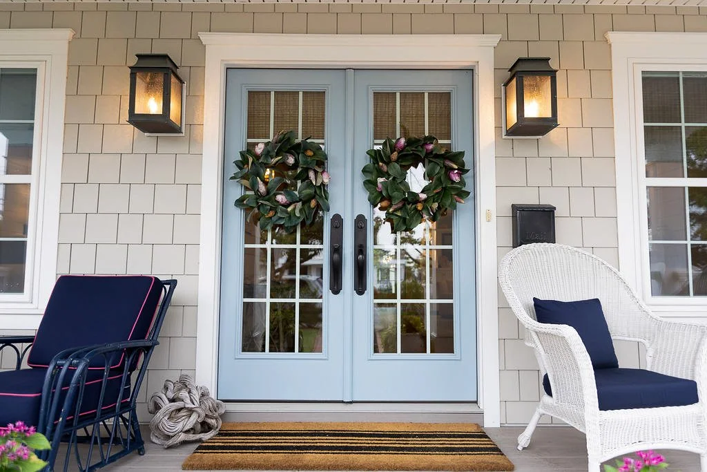 Front porch of a house with double glass doors decorated with two large wreaths. Flanking the doors are wall-mounted lanterns. To the left is a black outdoor chair with cushions, to the right is a white wicker chair with a pillow. A striped doormat i