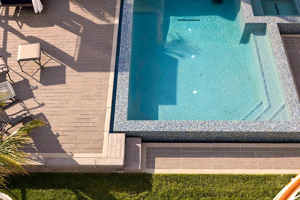 A swimming pool with clear blue water, surrounded by a tan and brown tiled deck, with patio chairs and a small table nearby.