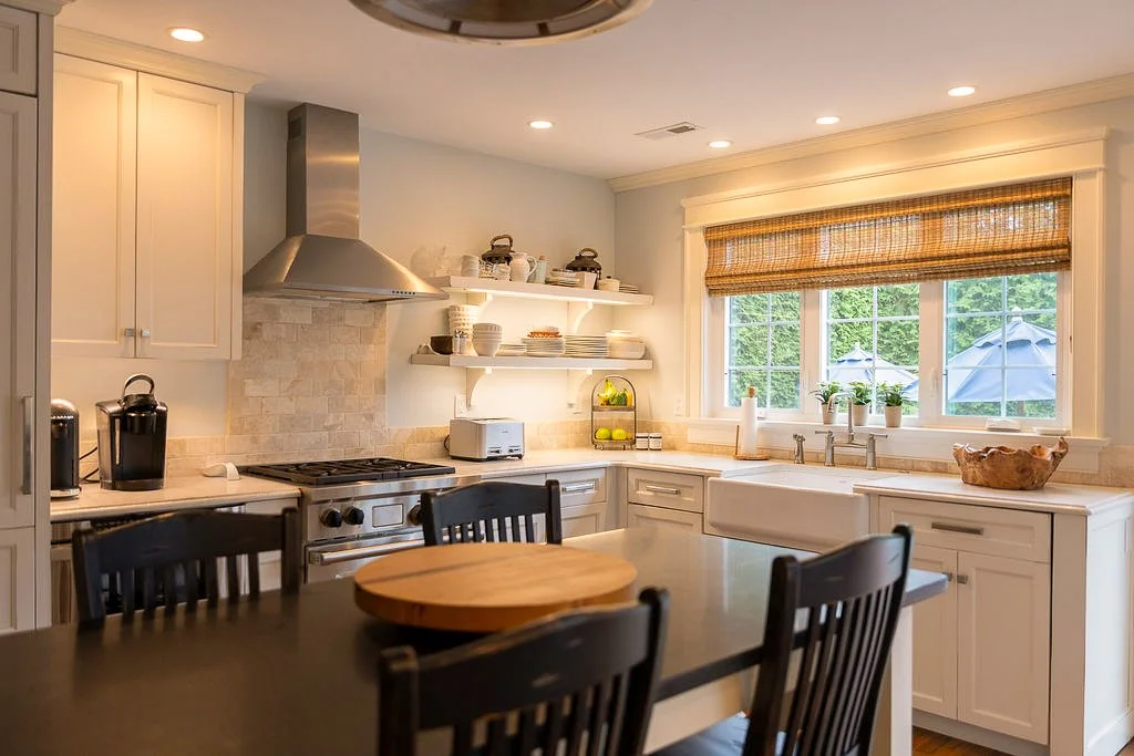 Bright kitchen with white cabinets, a large window with bamboo shades, open shelves with dishes, a stove with a stainless steel range hood, a farmhouse sink, and a round wooden table with black chairs.