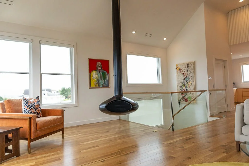 Modern living room with wooden flooring, orange sofa with patterned pillows, artwork on white walls, a black hanging fireplace, large windows, and a glass railing overlooking a staircase.