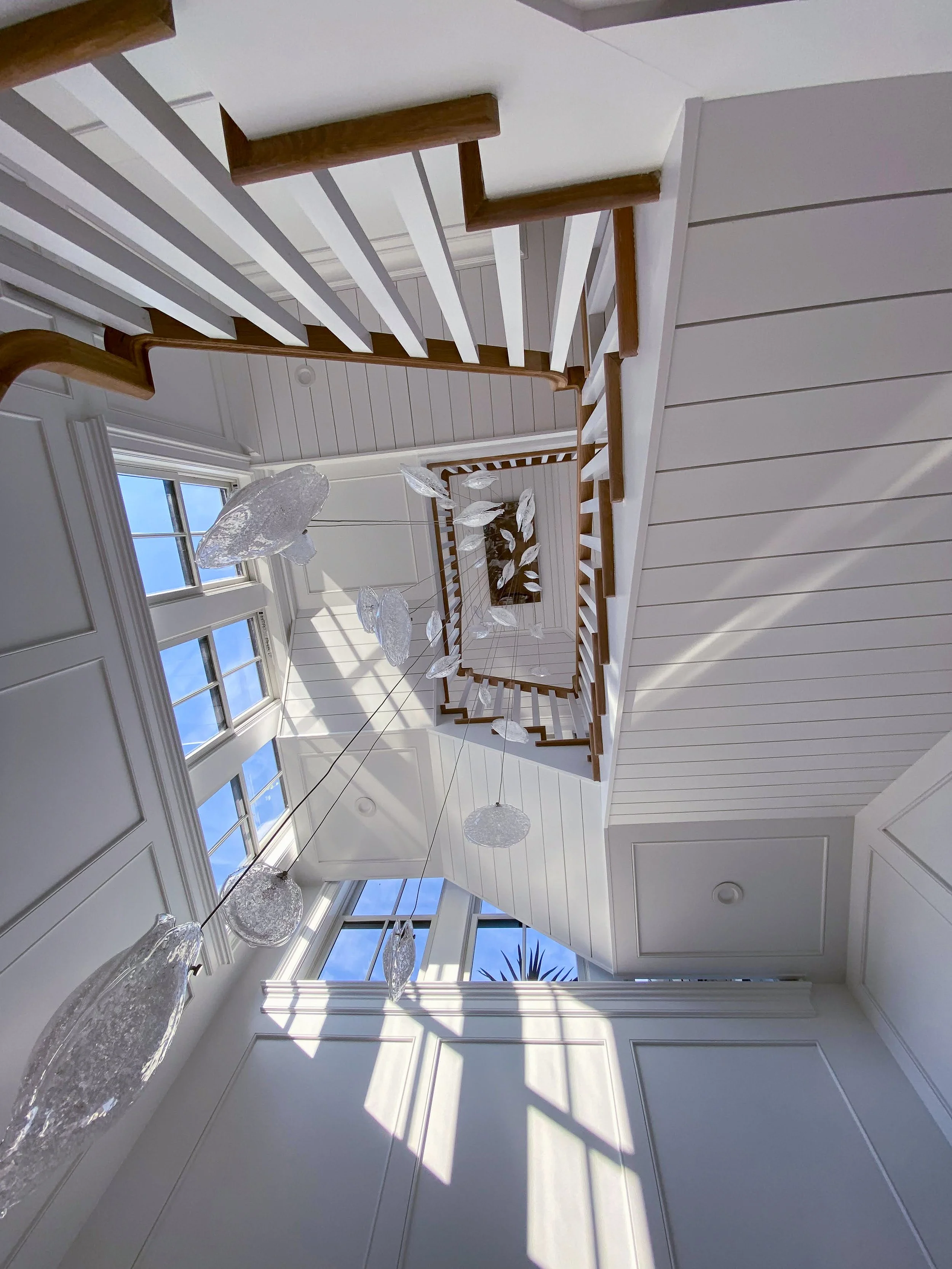 A view from the bottom of a staircase looking up, showing white walls with paneling, large windows letting in sunlight, and hanging glass light fixtures.