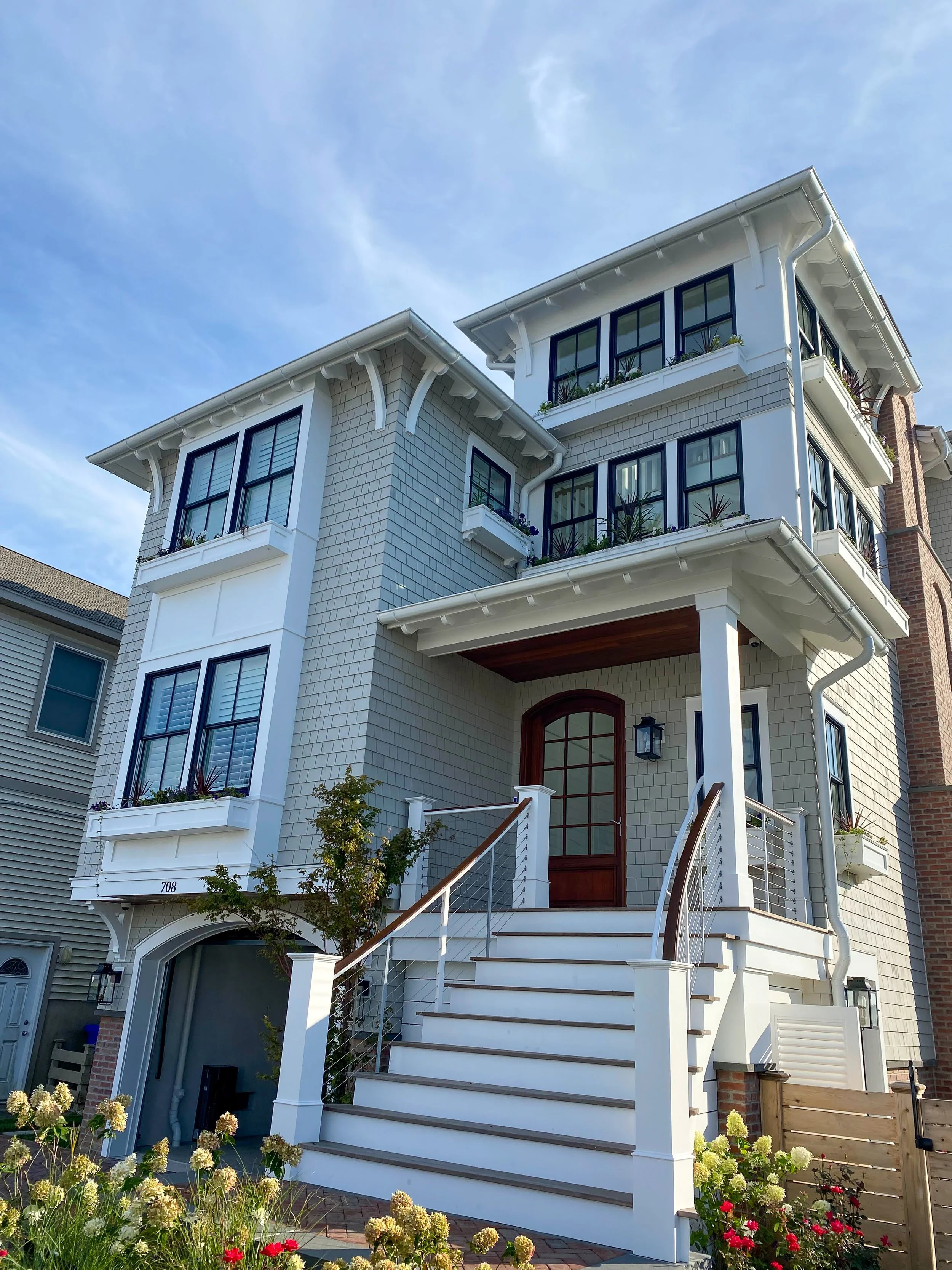 A multi-story house with white exterior, large windows, a front staircase, and decorative flowers in the yard under a blue sky.
