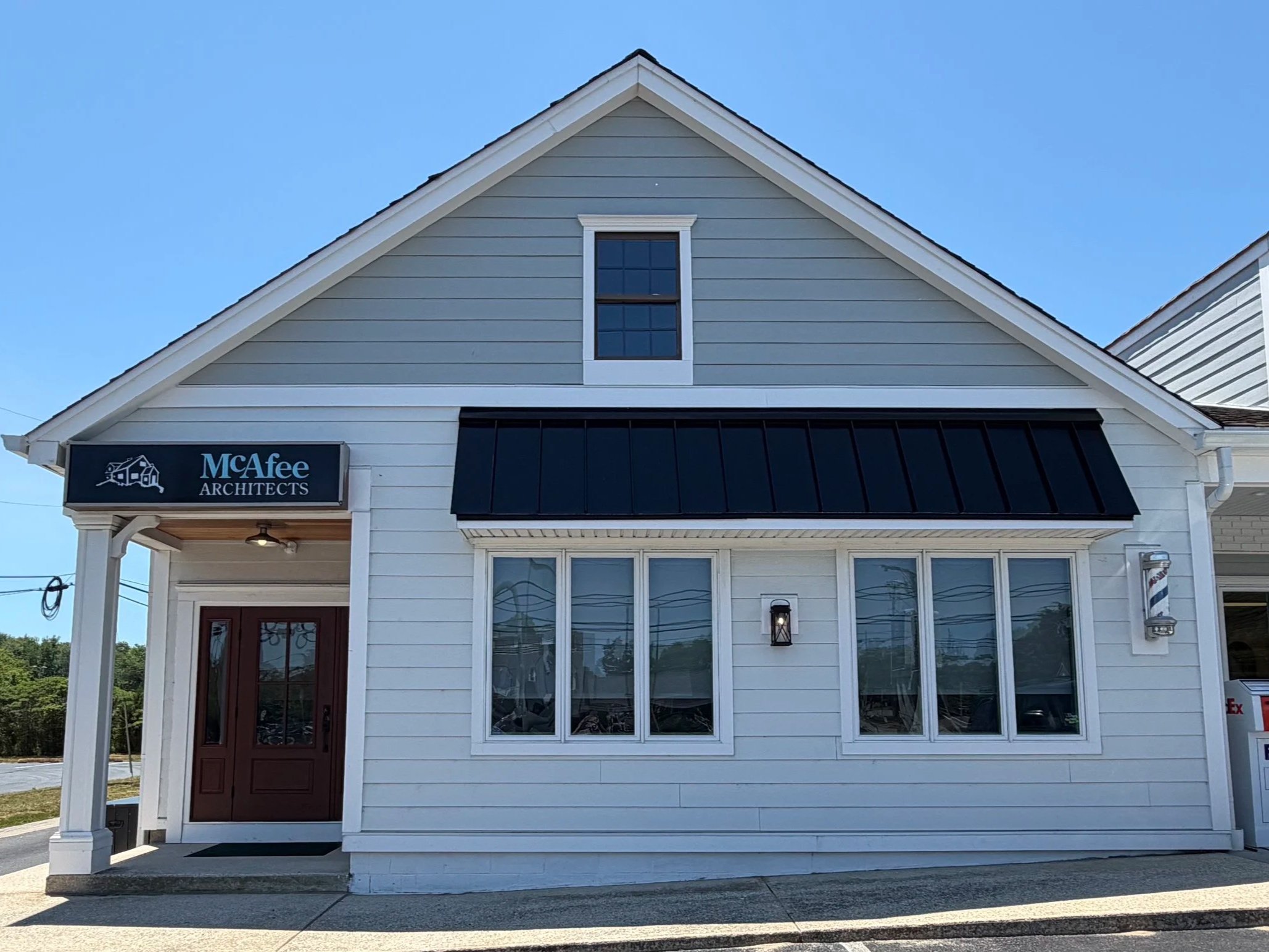 Front view of a light blue house with white trim, a gabled roof, and a sign that reads "McAfee Architects" on the front. The house has a small porch with a wooden door and large windows in the lower part. There is a black metal awning above the windows and a small outdoor light fixture on the wall. The sky is clear and blue.