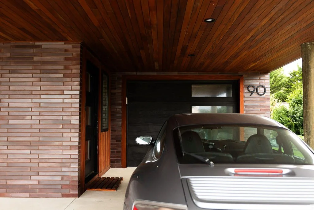 Modern brick house with a garage, black garage door, and a vintage gray car parked outside. The house has house number 90, a wooden front door, and a wood-paneled porch ceiling.