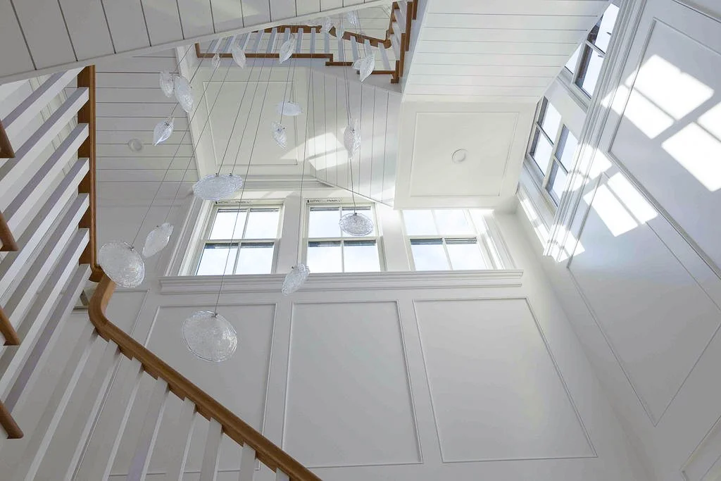 Inside view of a bright, white staircase area with large windows, wall paneling, and hanging glass light fixtures.