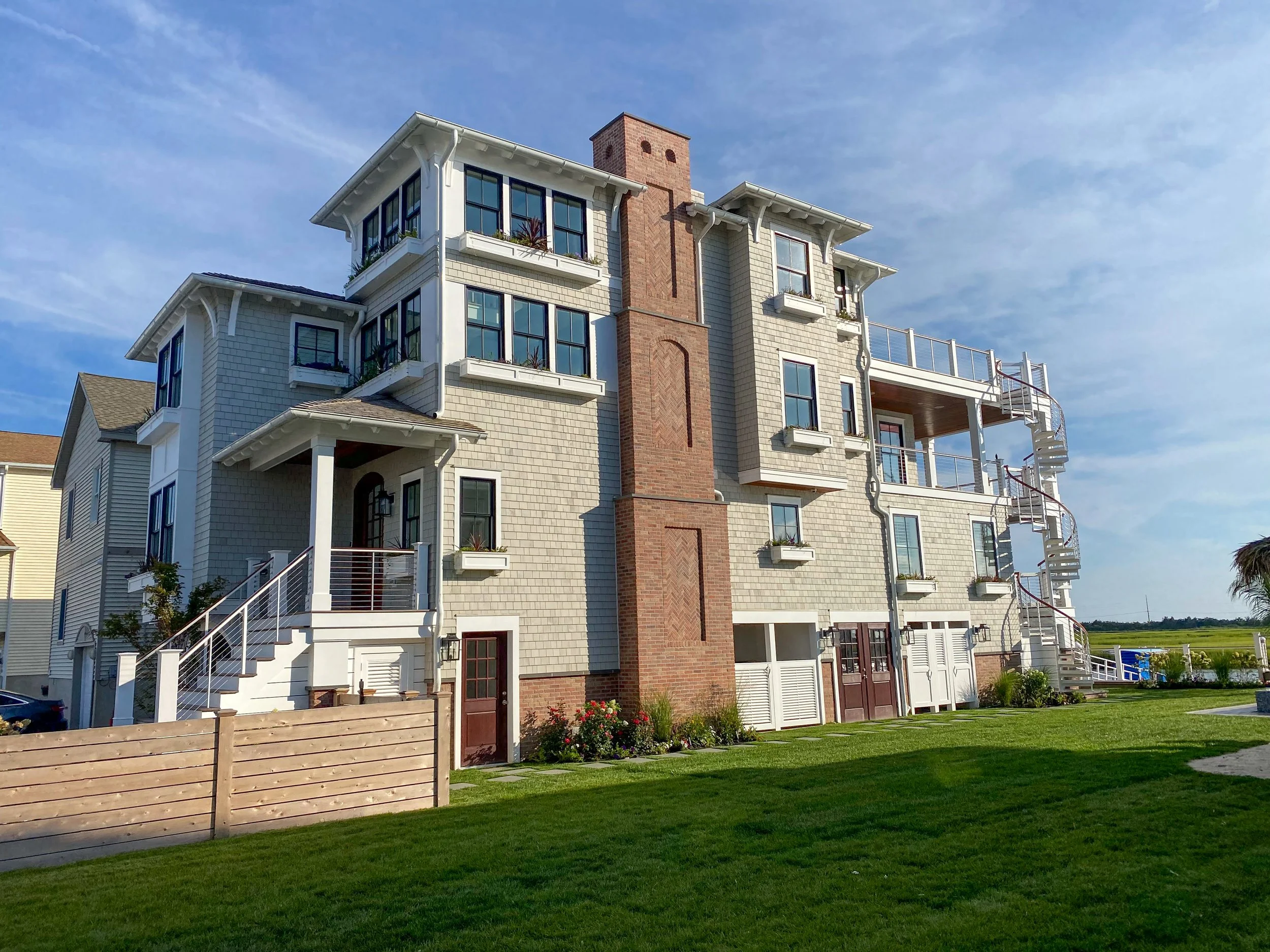 A multi-story Bayfront home with a mix of white siding and brick exterior, many windows, a spiral exterior staircase, a wooden fence, green lawn, and a partly cloudy blue sky.