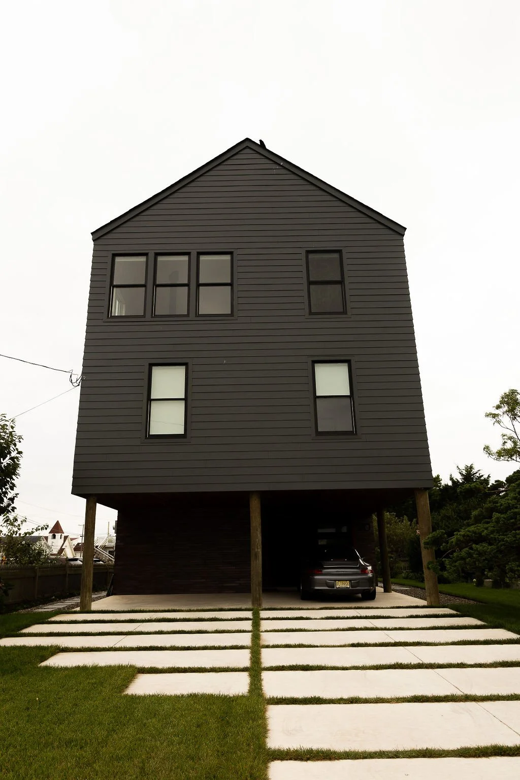 A modern three-story house with black siding, large windows, and an elevated parking area beneath. The driveway has concrete slabs with grass in between.