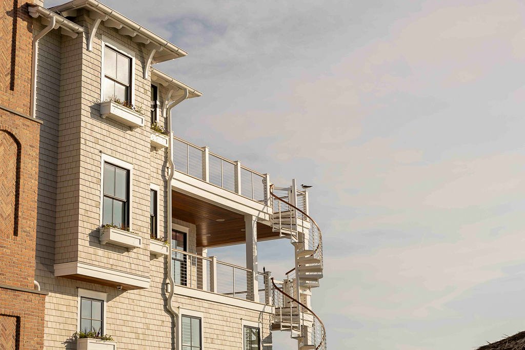 An exterior view of a Bayfront multi-story house with balconies, spiral staircase, and beige siding, against a partly cloudy sky.