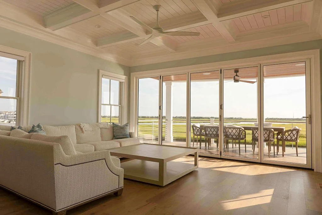Living room with large glass sliding doors opening to a patio with a dining table and chairs, overlooking a grassy landscape.