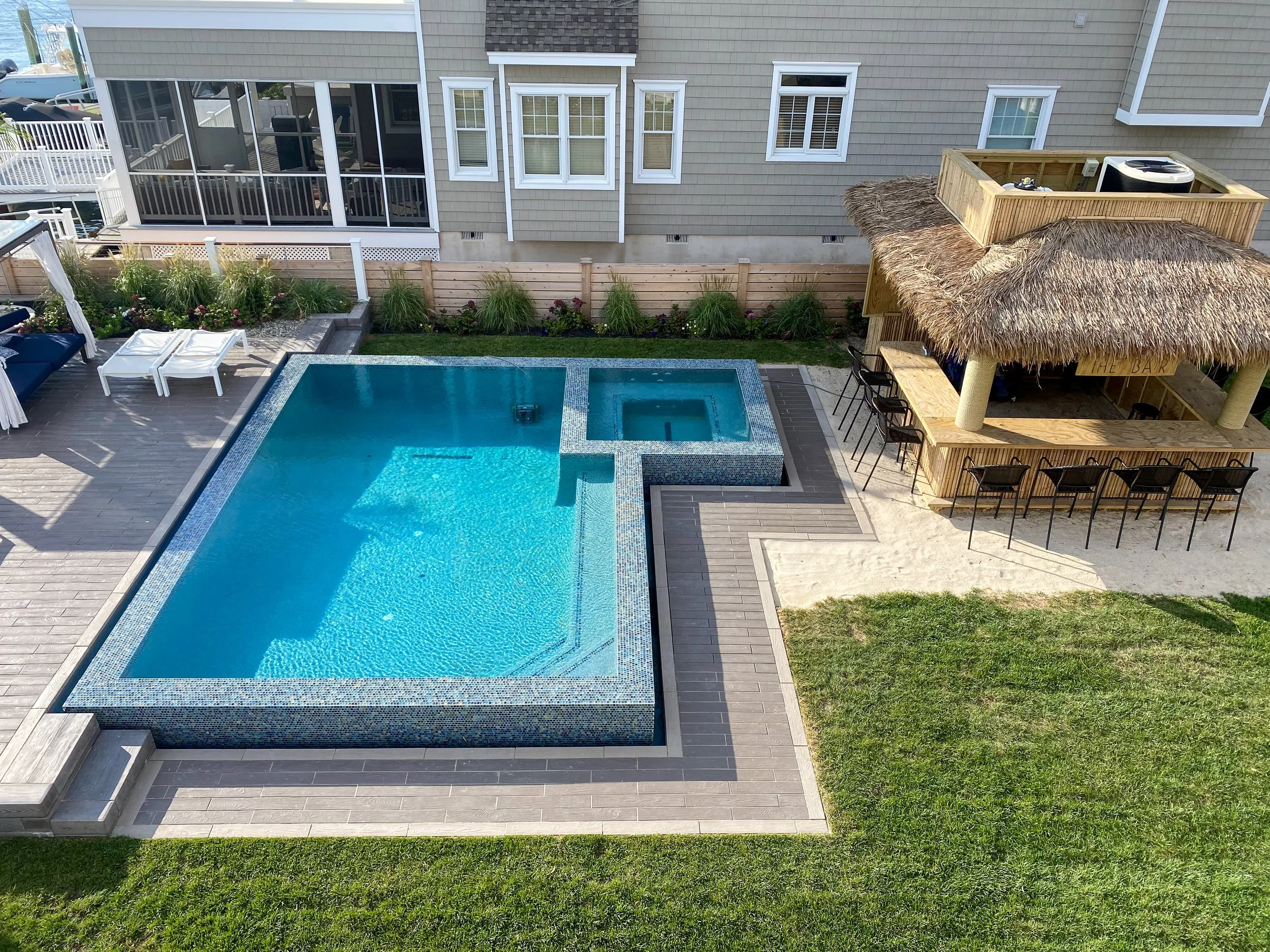 A backyard with a swimming pool, a hot tub, an outdoor bar with seating, a fenced garden, and an upper deck with chairs, all next to a house with multiple windows.