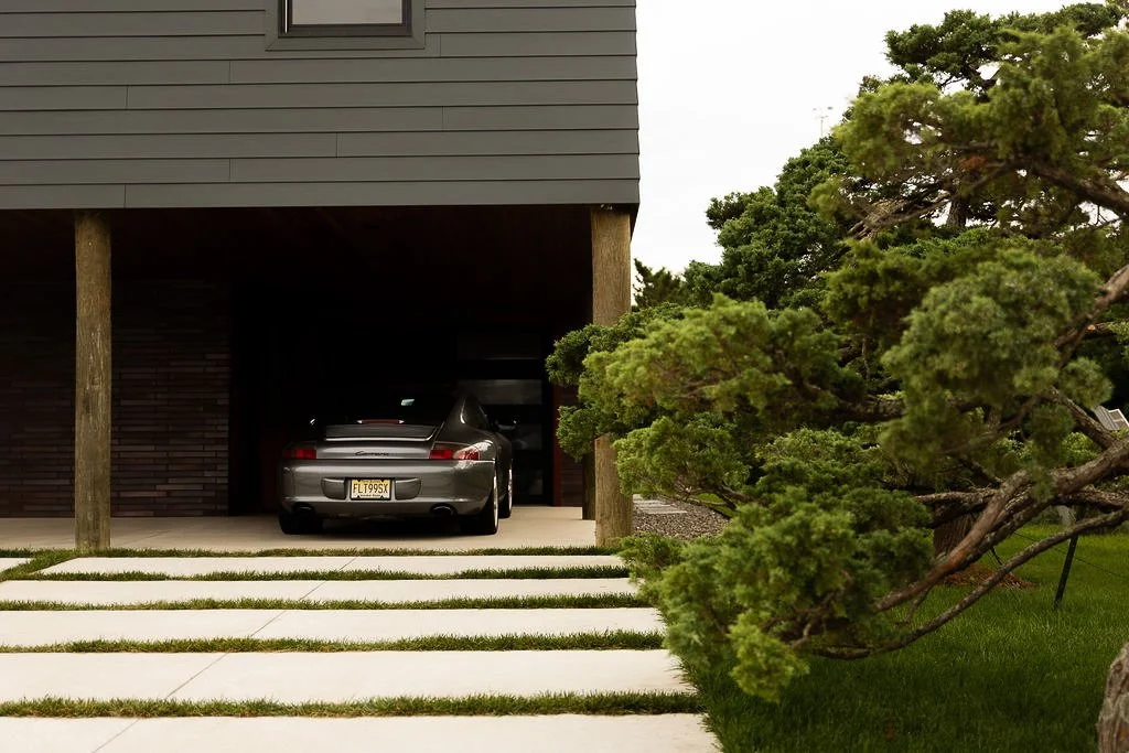 Silver sports car parked in a garage of a modern house with grey exterior, wooden pillars, and a landscaped yard with large green trees and grass.