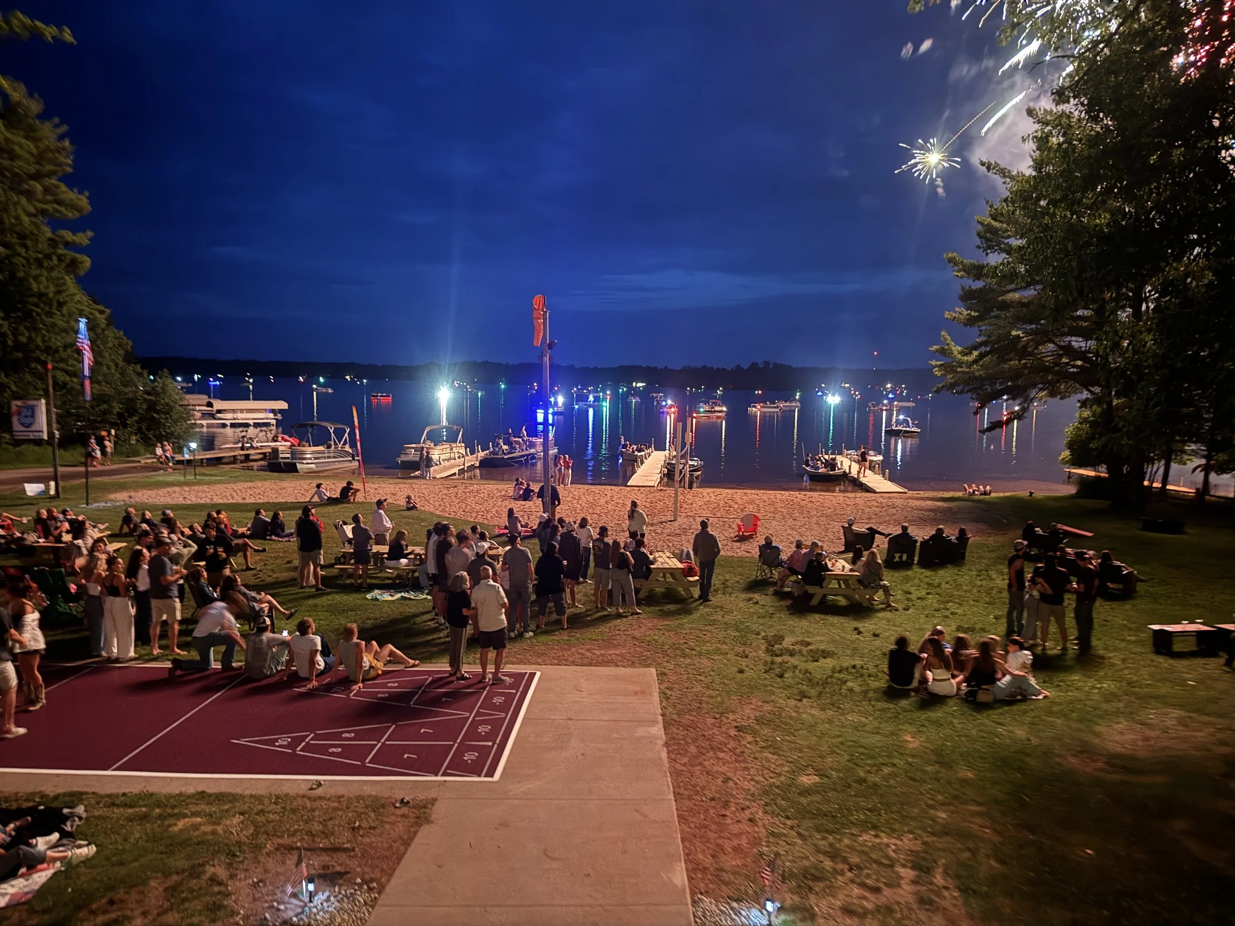 People gathered at a waterfront park during fireworks at night, with boats on the water and trees lining the area.