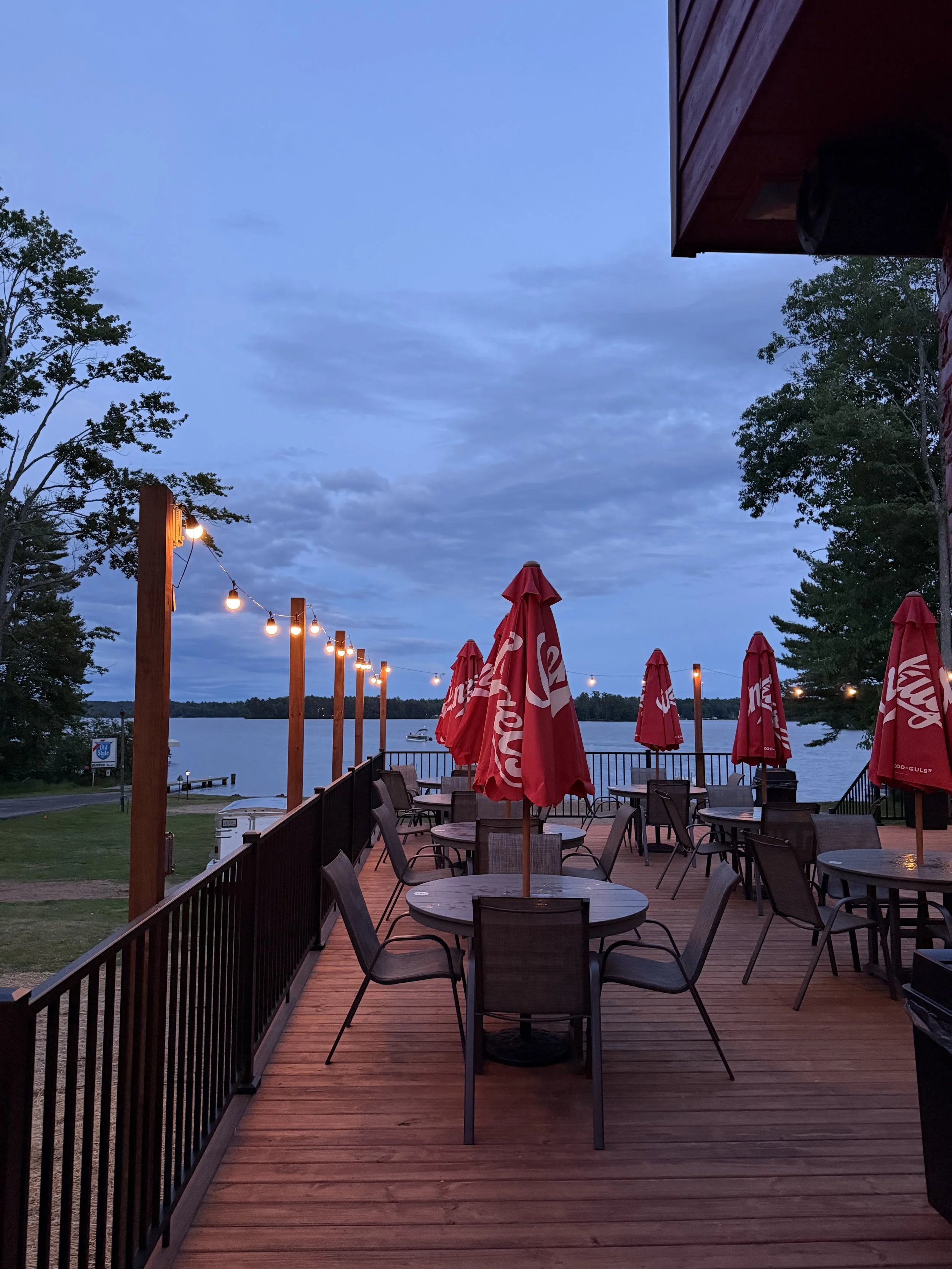 Outdoor patio with wooden flooring, round tables, red umbrellas, string lights, and trees near a body of water during dusk.