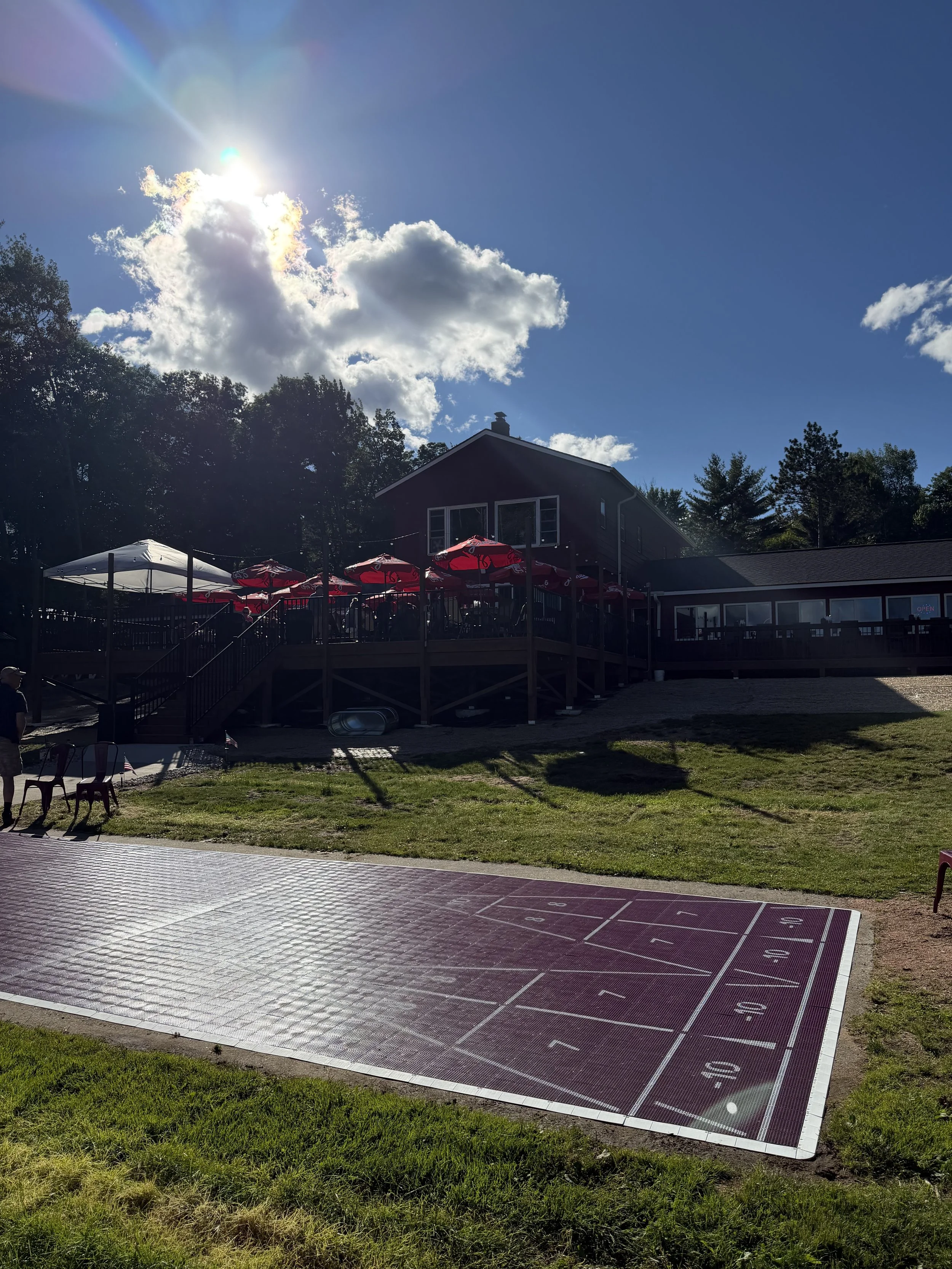 An outdoor scene featuring a large, empty shuffleboard court with numbered sections, a grassy area, a building with an outdoor patio with umbrellas, and a clear blue sky with some clouds and the sun shining brightly.