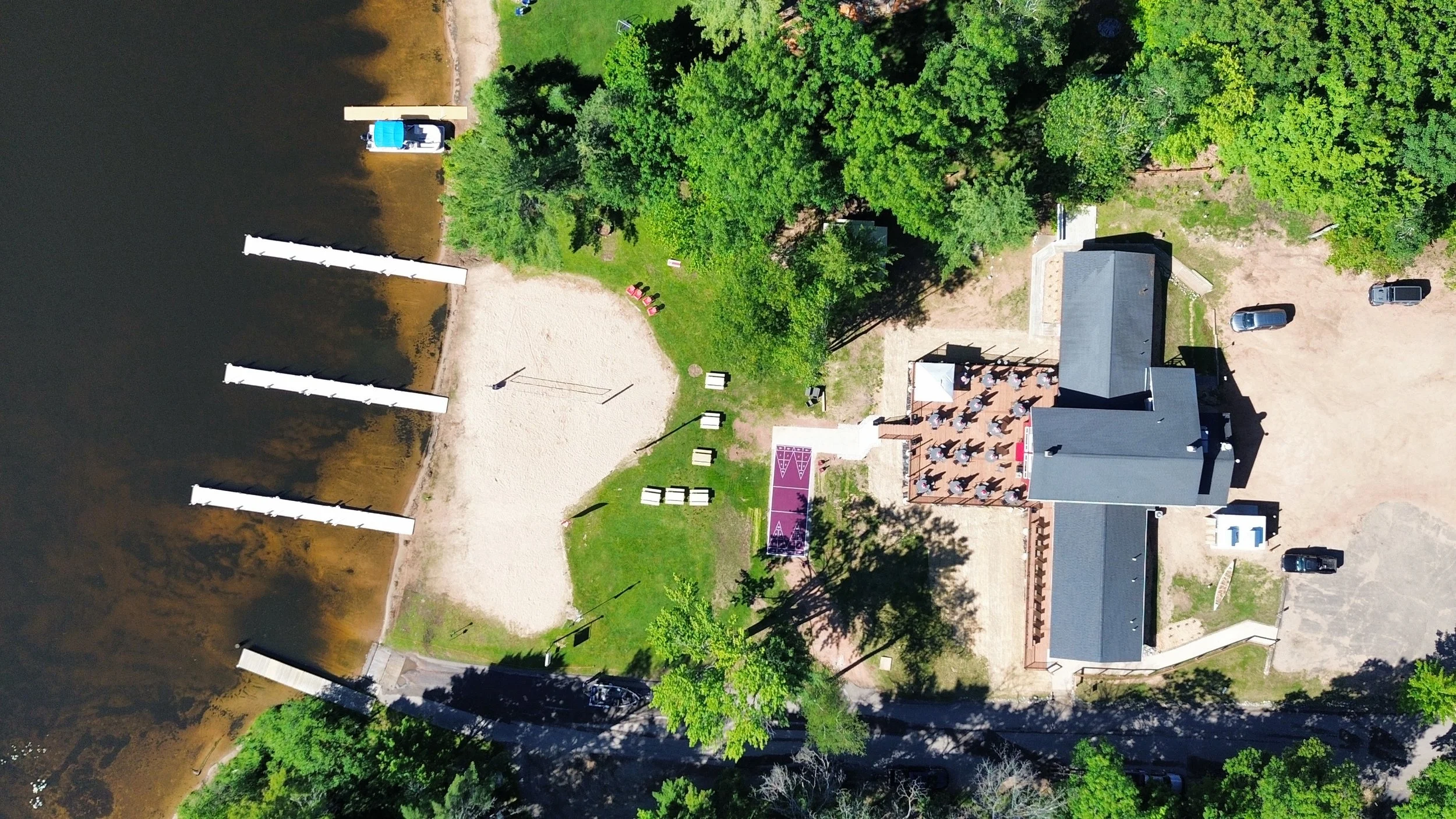 An aerial view of a lakeside area featuring a building, parking area with cars, a sand volleyball court, a grassy picnic area with tables, a boat dock with boats, and lush green trees surrounding the area.