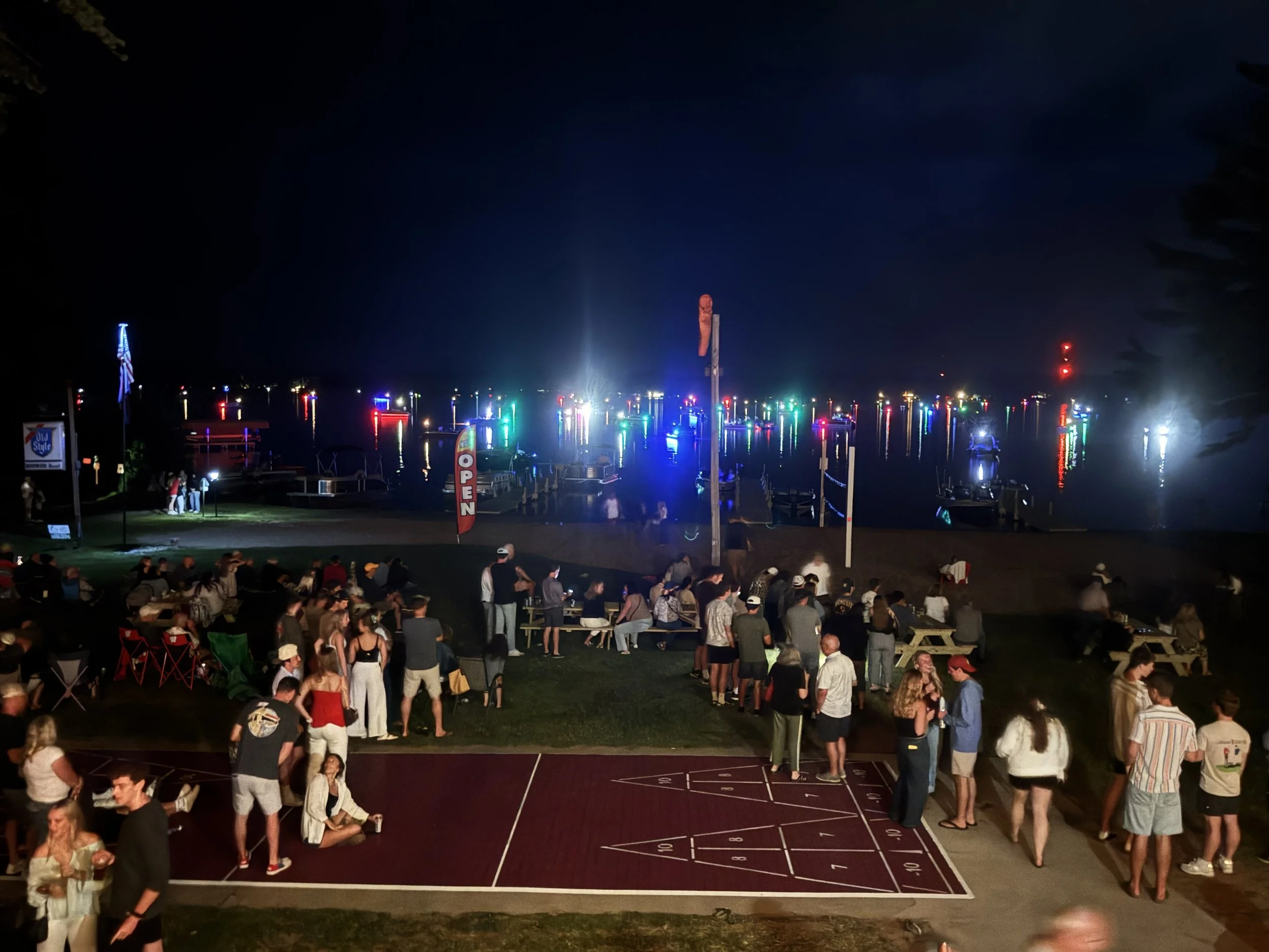 Night scene at a marina with boats and colorful lights reflecting on the water, crowd of people on the grass and a basketball court in the foreground.