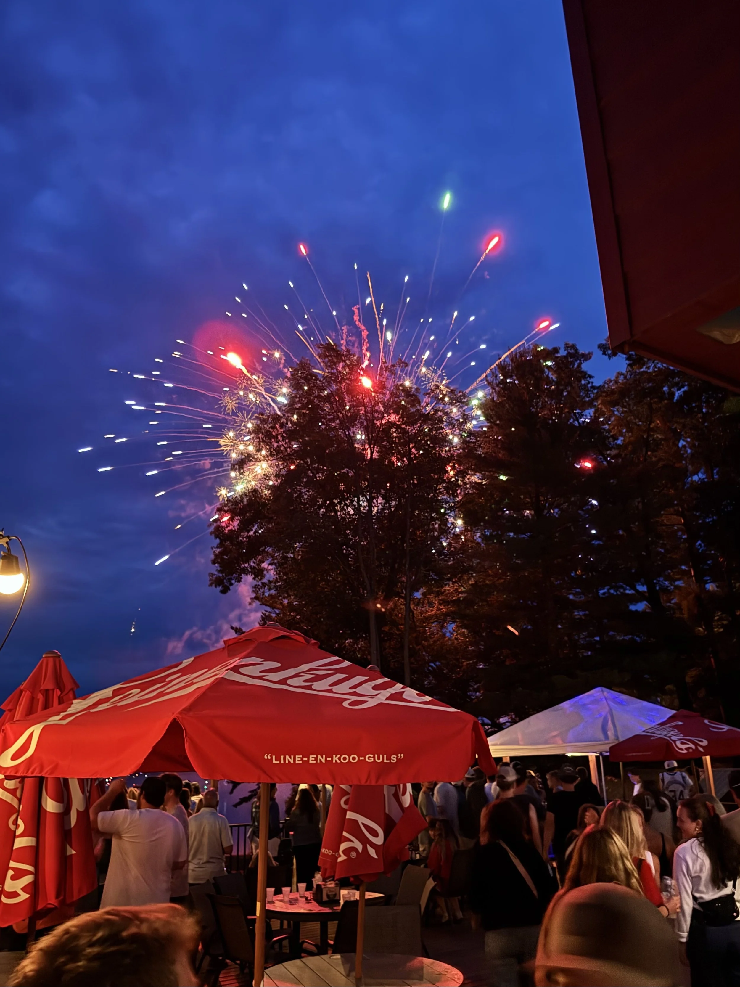 People gathered outdoors at a nighttime event with fireworks lighting up the sky, large red umbrellas with Coca-Cola branding, and various tents or canopies.
