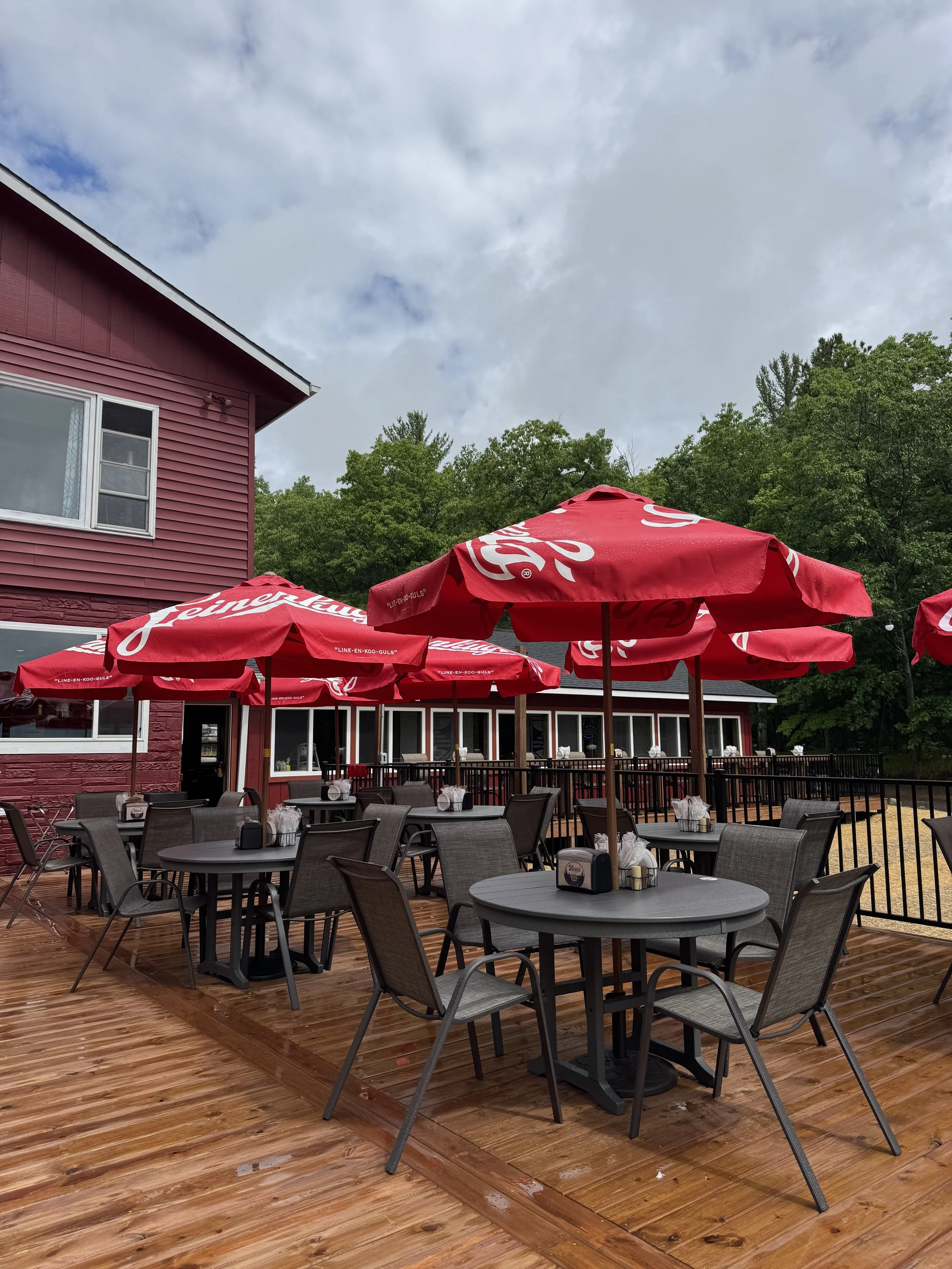 Outdoor patio area with multiple tables and chairs, red umbrellas with white branding, adjacent to a red building with a white half-moon window and surrounded by green trees, on a wooden deck under a partly cloudy sky.