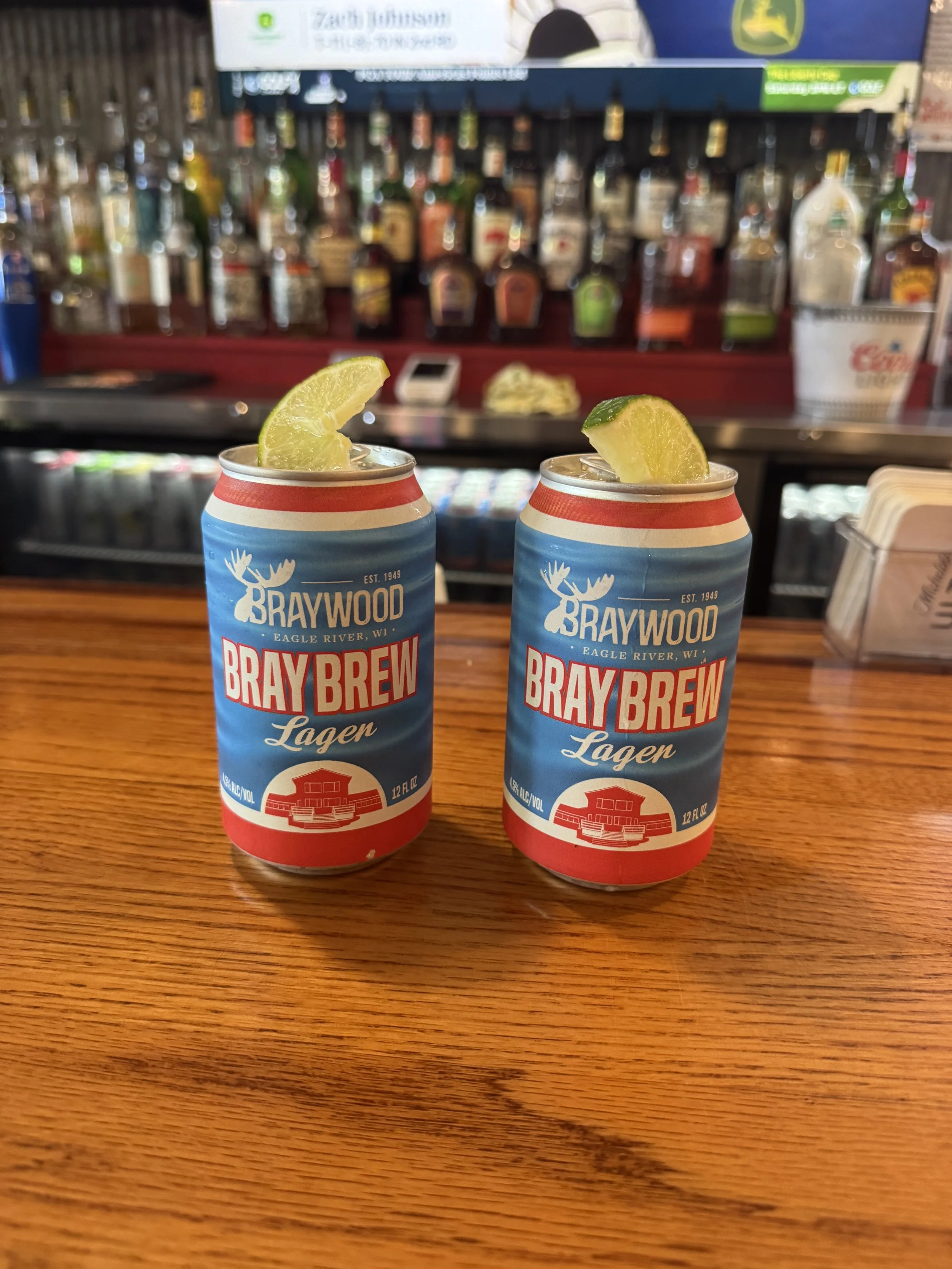Two cans of BrayBrew Lager with lemon wedges on top, placed on a wooden surface in a bar setting with liquor bottles and bar equipment in the background.