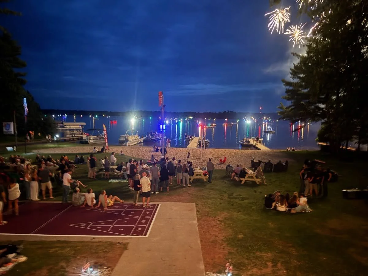 A nighttime scene at a waterfront with fireworks in the sky. People are gathered on the grass and picnic tables watching the fireworks and enjoying the event. Boats are docked on the water with colorful lights reflecting on the surface.